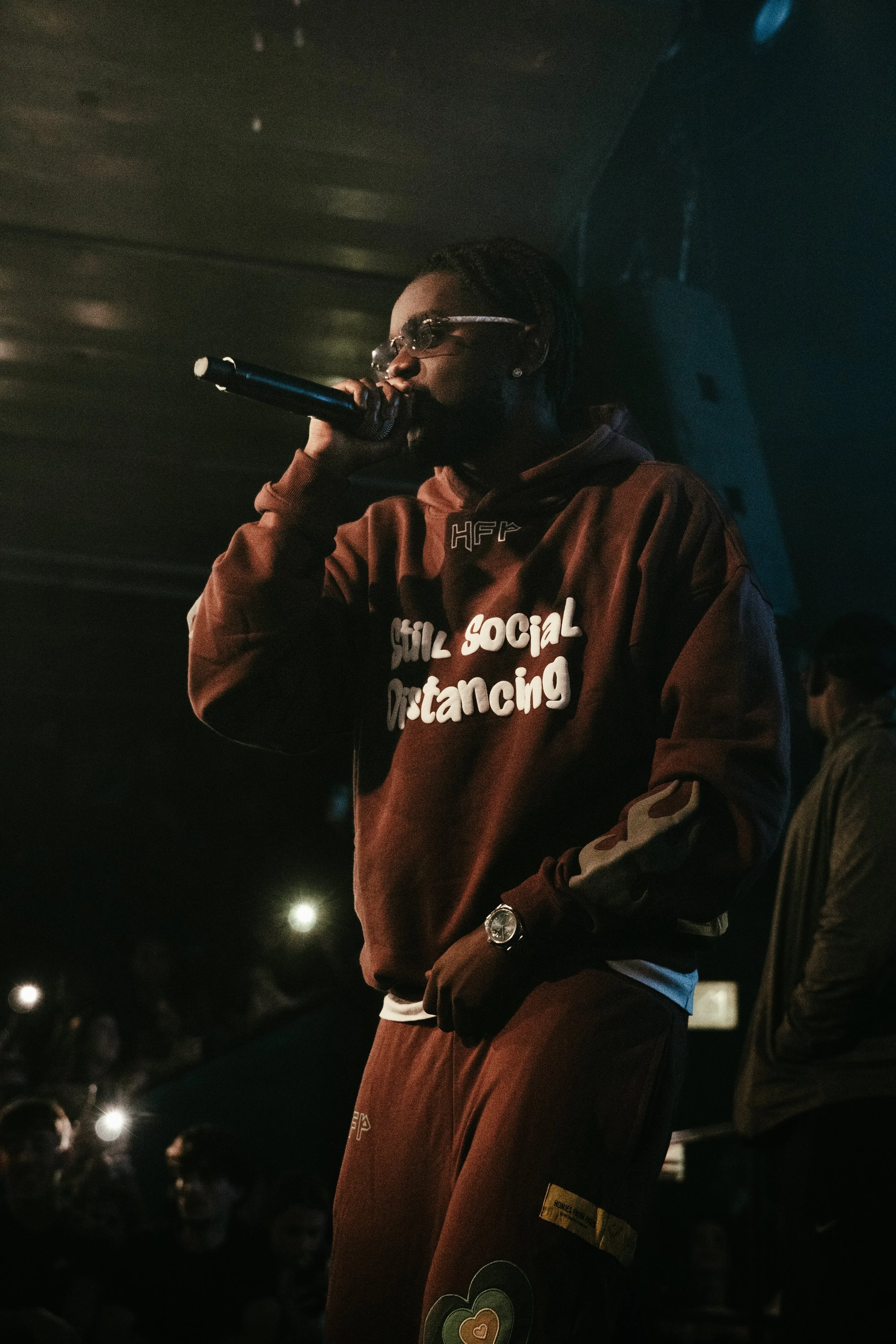 Une personne portant un sweat à capuche marron avec le texte 'social distancing' à l'avant, tenant un microphone lors d'une performance dans une ambiance sombre et animée.