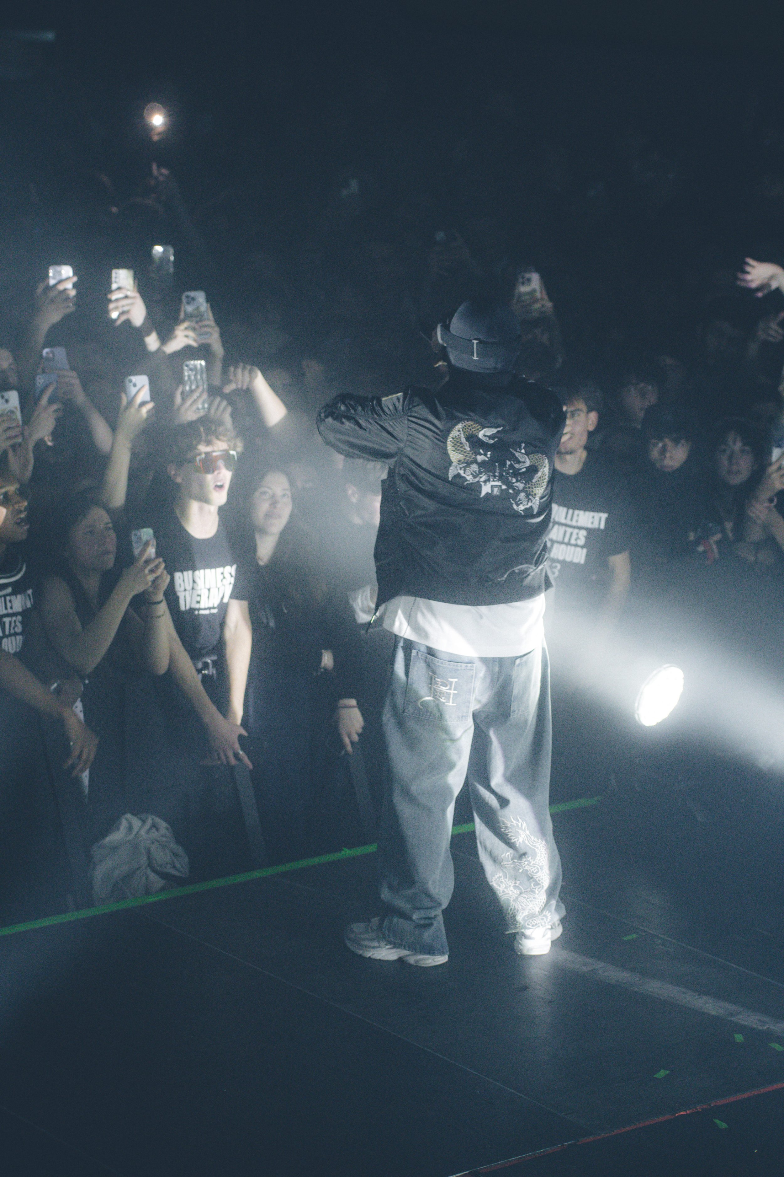 Un artiste sur scène avec un microphone, entouré d'un public nombreux qui l'enregistre avec leurs téléphones, dans une ambiance sombre et éclairée.