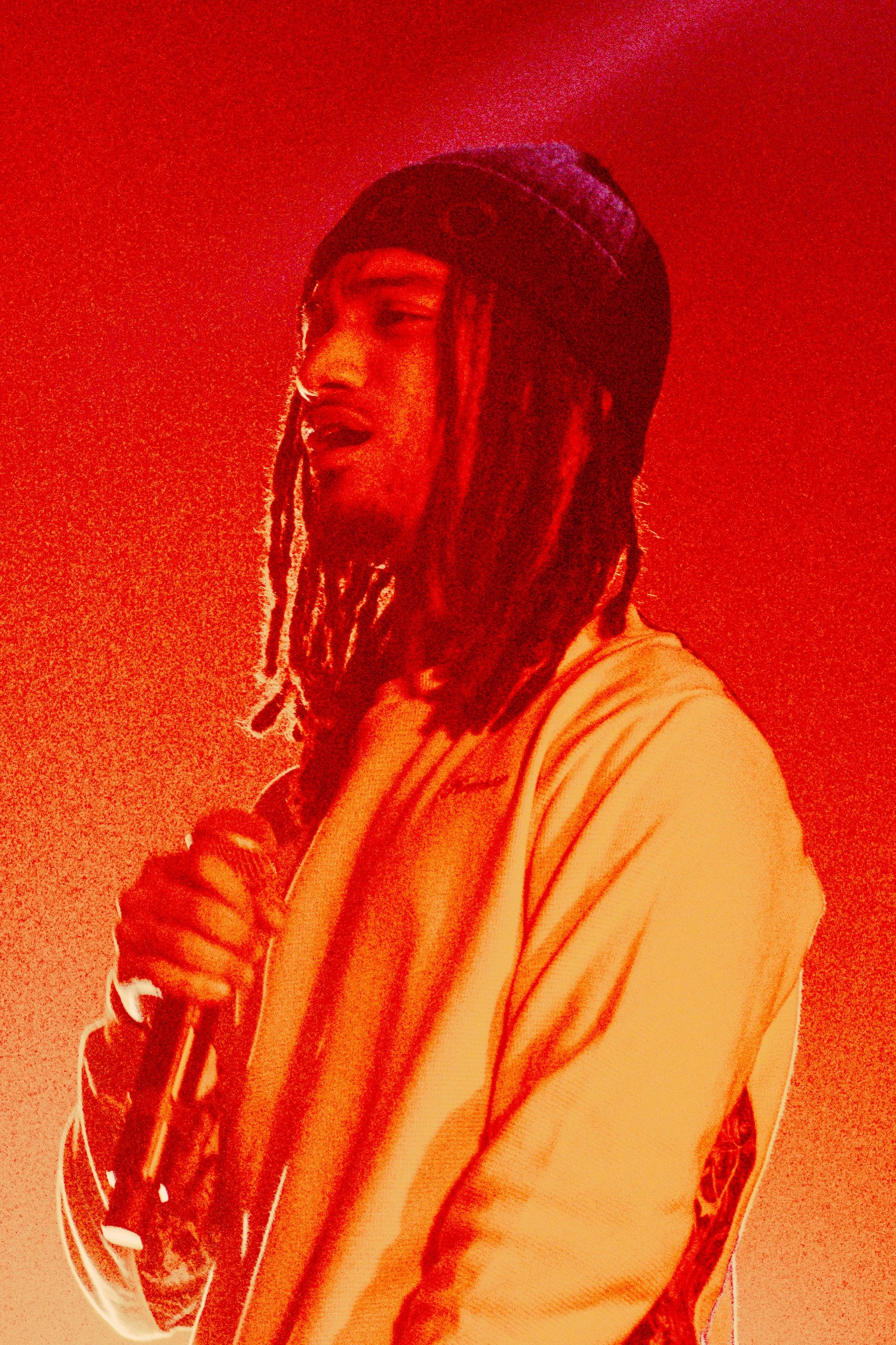 Un homme avec des dreadlocks porte un casque et une veste blanche, tenant un microphone, sur un fond rouge.