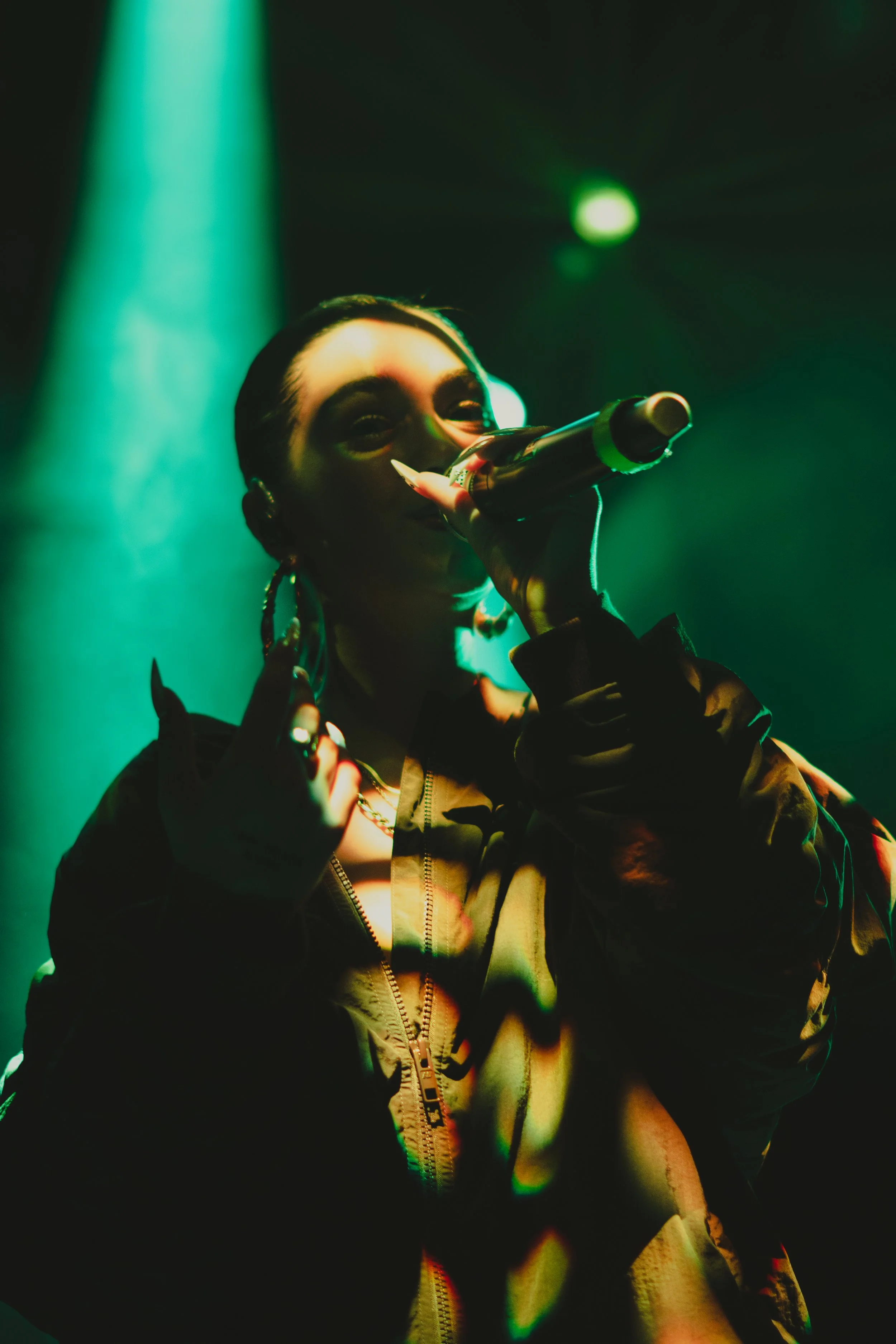 Une femme qui chante ou parle dans un microphone lors d'une performance, sous un éclairage vert et sombre.