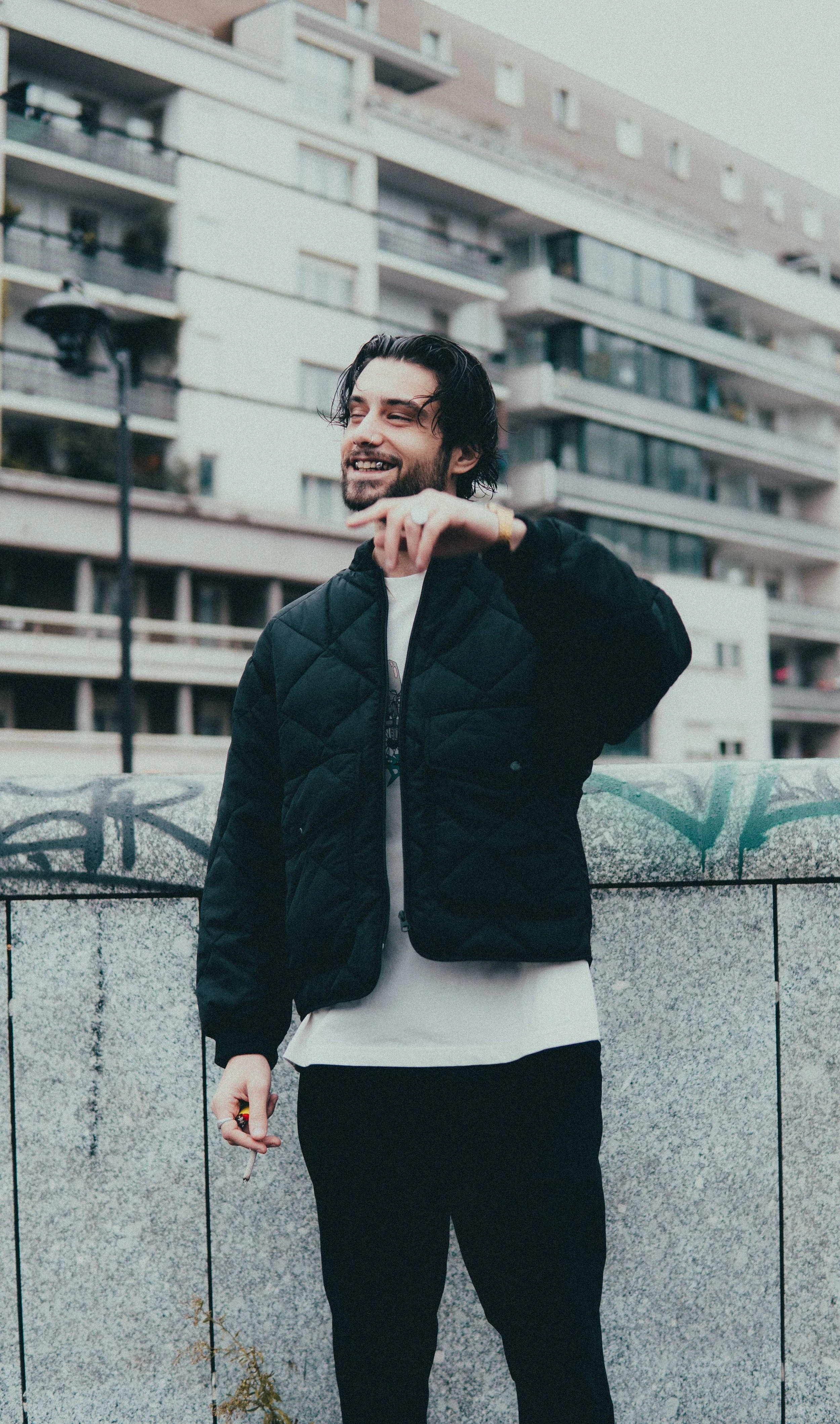 Jeune homme souriant avec des cheveux longs et une barbe, portant une veste noire et un t-shirt blanc, debout devant un mur en pierre avec des graffitis, dans un environnement urbain avec des immeubles en arrière-plan.