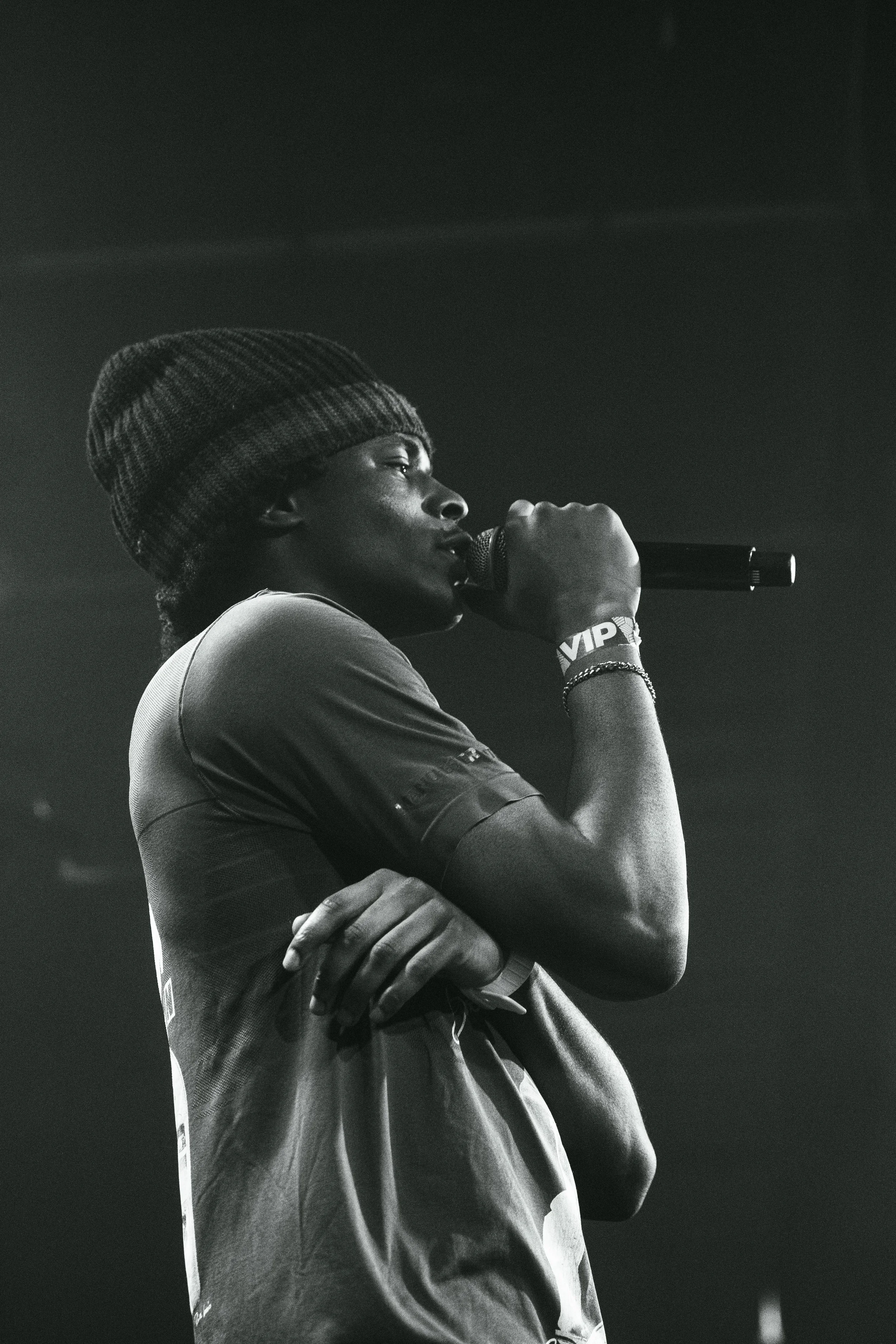 Une femme noire porte un bonnet, tient un microphone et semble chanter ou parler lors d'une performance, en noir et blanc.