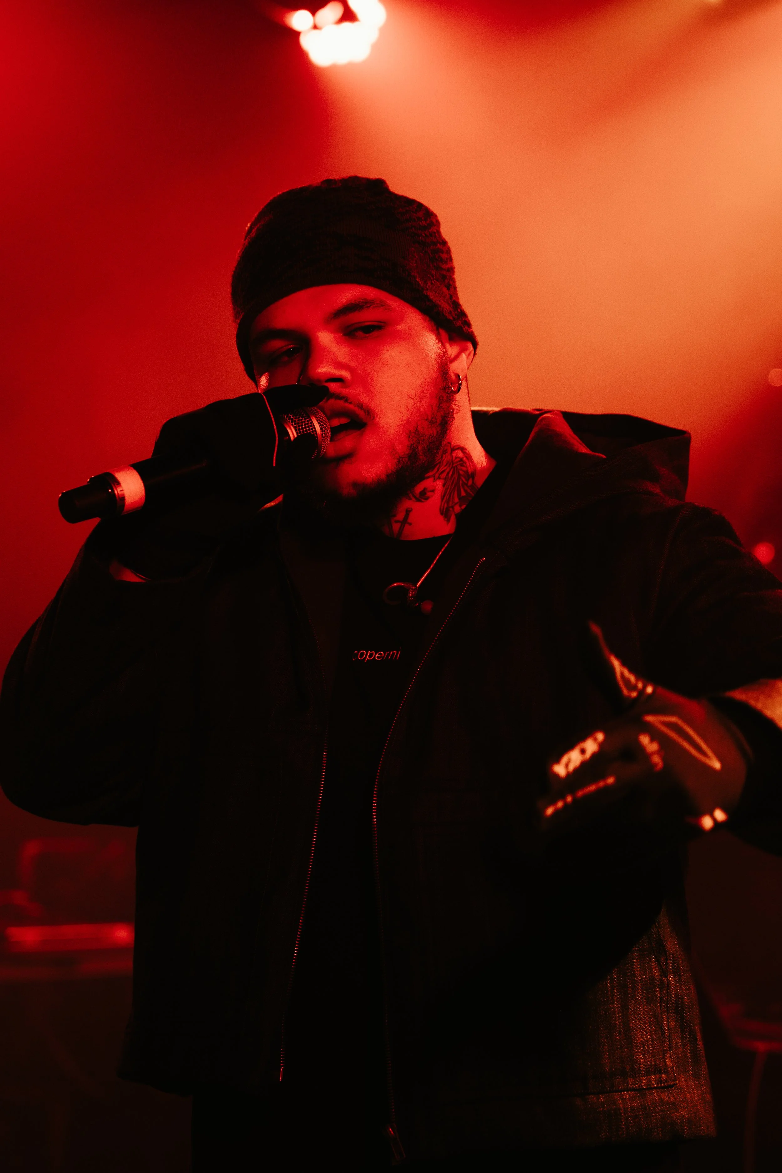 Un jeune homme avec un bonnet noir, des tatouages au cou, tenant un microphone en scène avec un éclairage rouge intense.