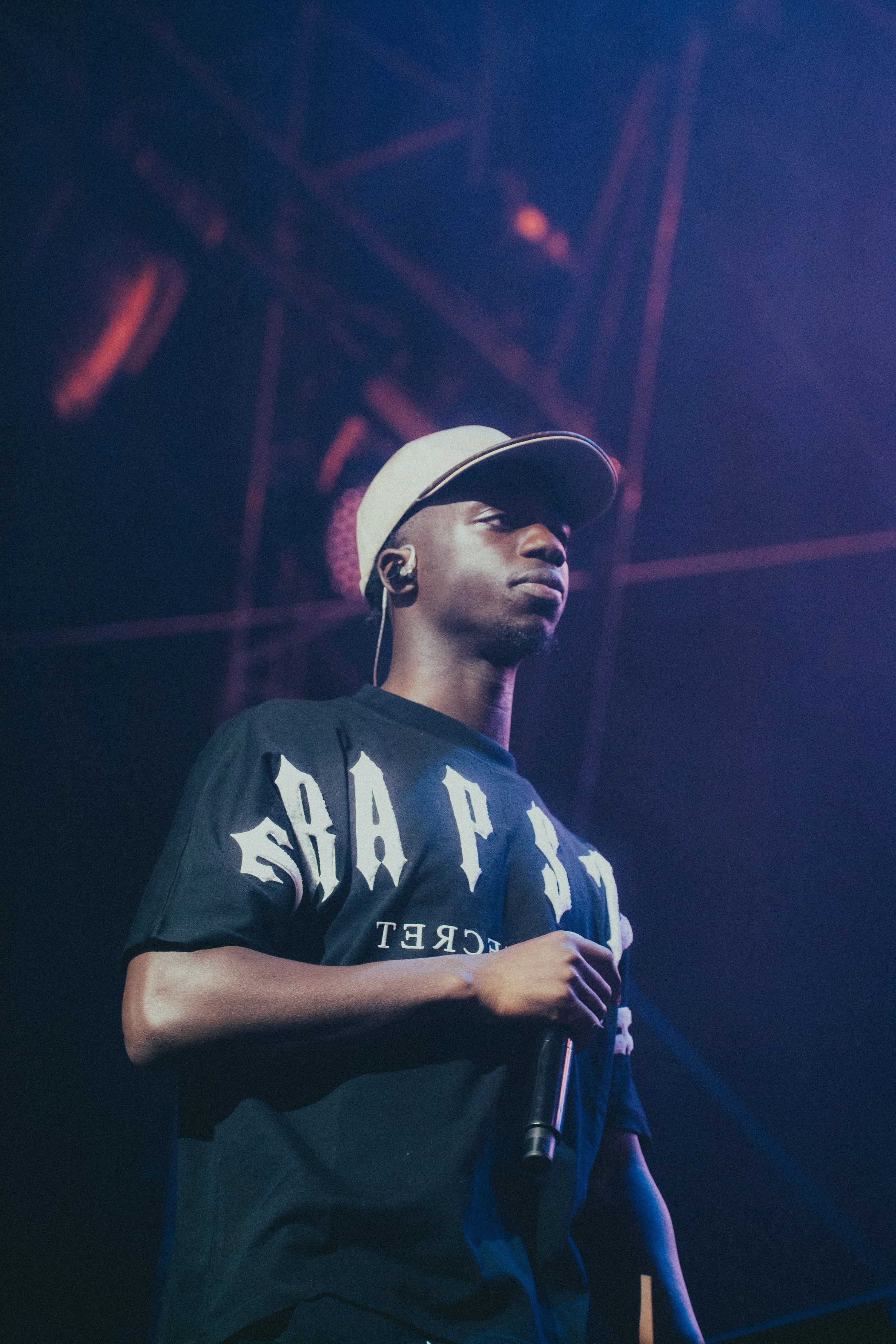Un jeune homme porte un t-shirt noir avec l'inscription 'PARIS' et un chapeau beige, tenant un micro lors d'une performance sur scène, sous un éclairage violet