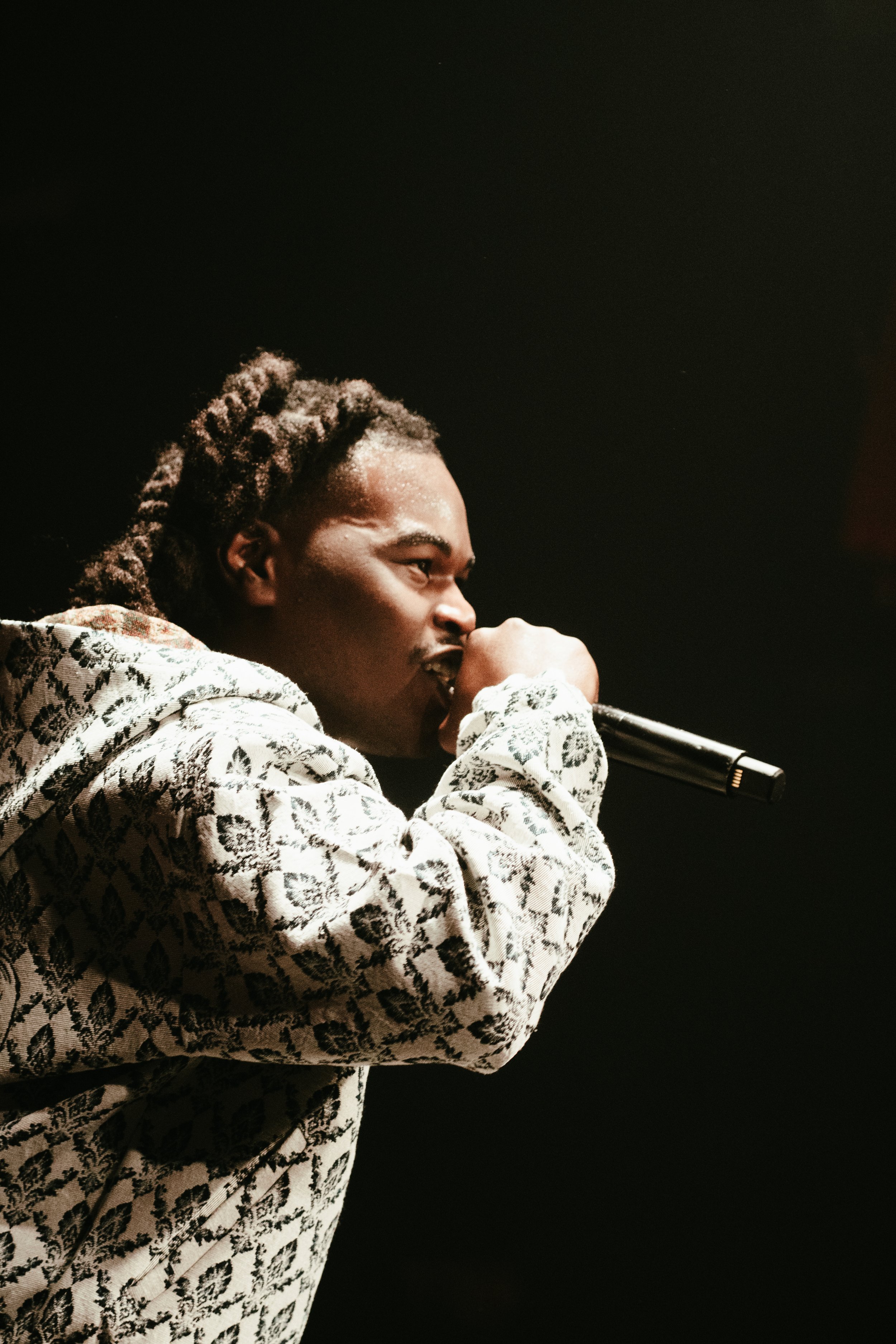 Un homme noir avec des cheveux en dreadlocks, portant un sweat à motif léopard, tient un micro près de sa bouche, en train de chanter ou de parler, dans un environnement sombre.