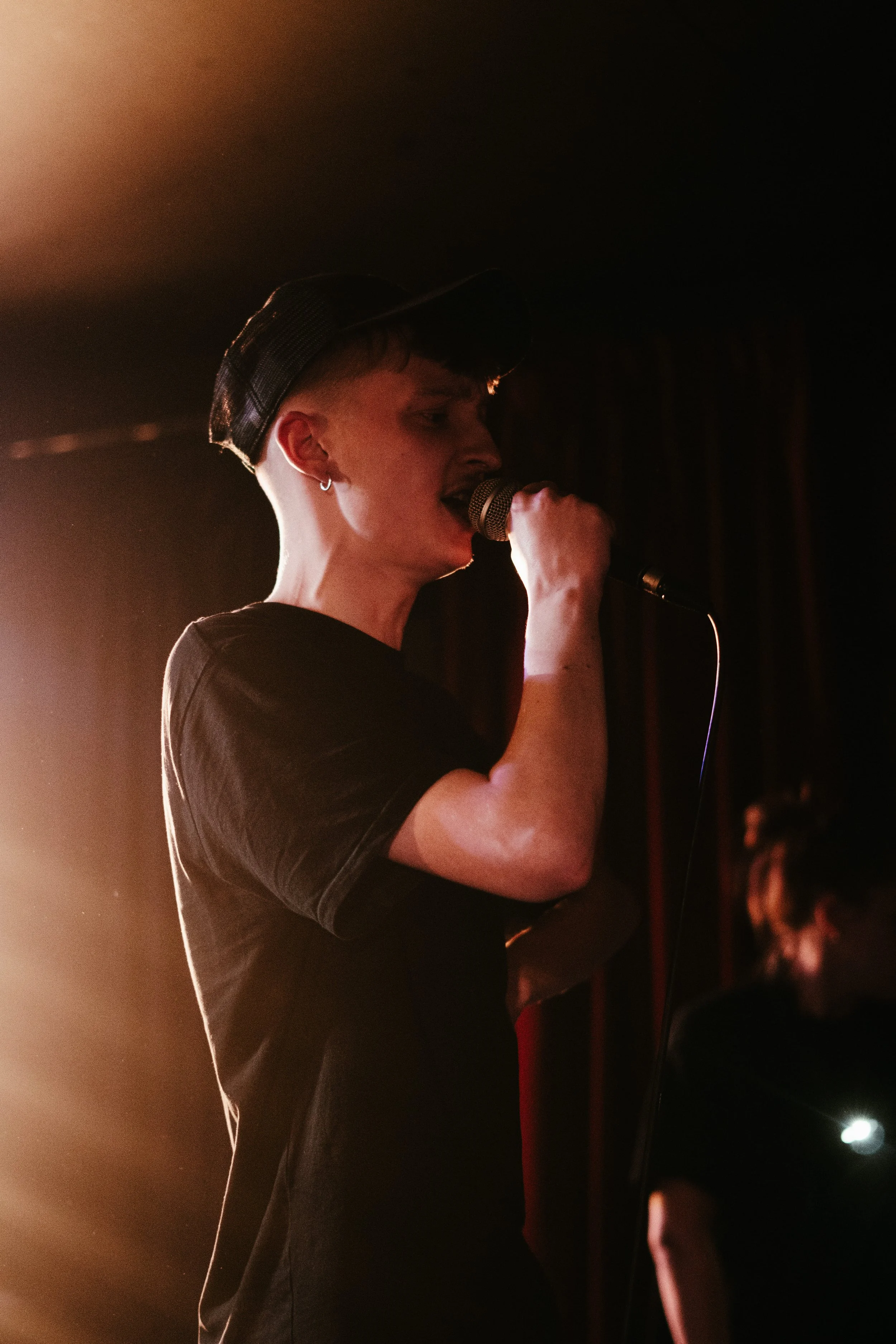 Une personne chante dans un microphone sur scène, éclairée par un éclairage chaud, portant un T-shirt noir et un chapeau noir en maille, avec un fond sombre.