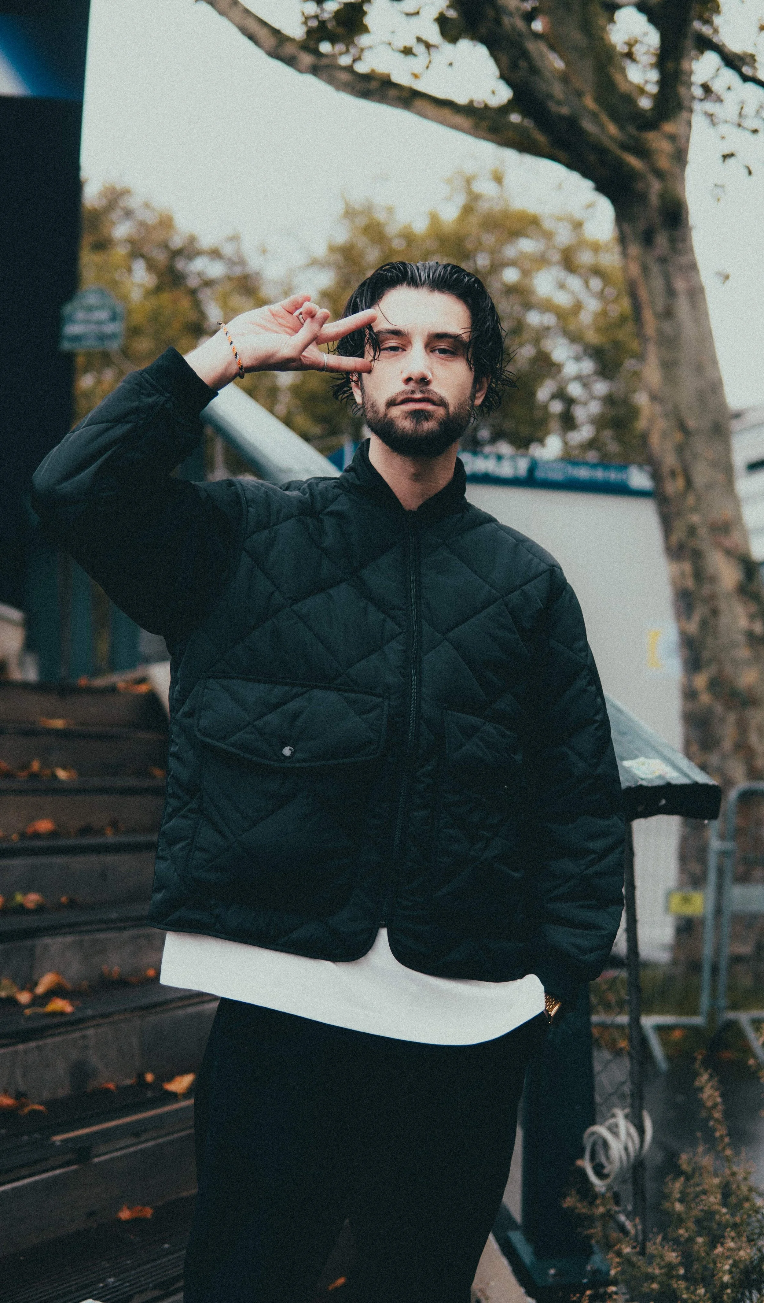 Un jeune homme pose en faisant un signe de paix avec la main, portant une veste noire et se trouvant à l'extérieur, près d'un arbre et d'une échelle.