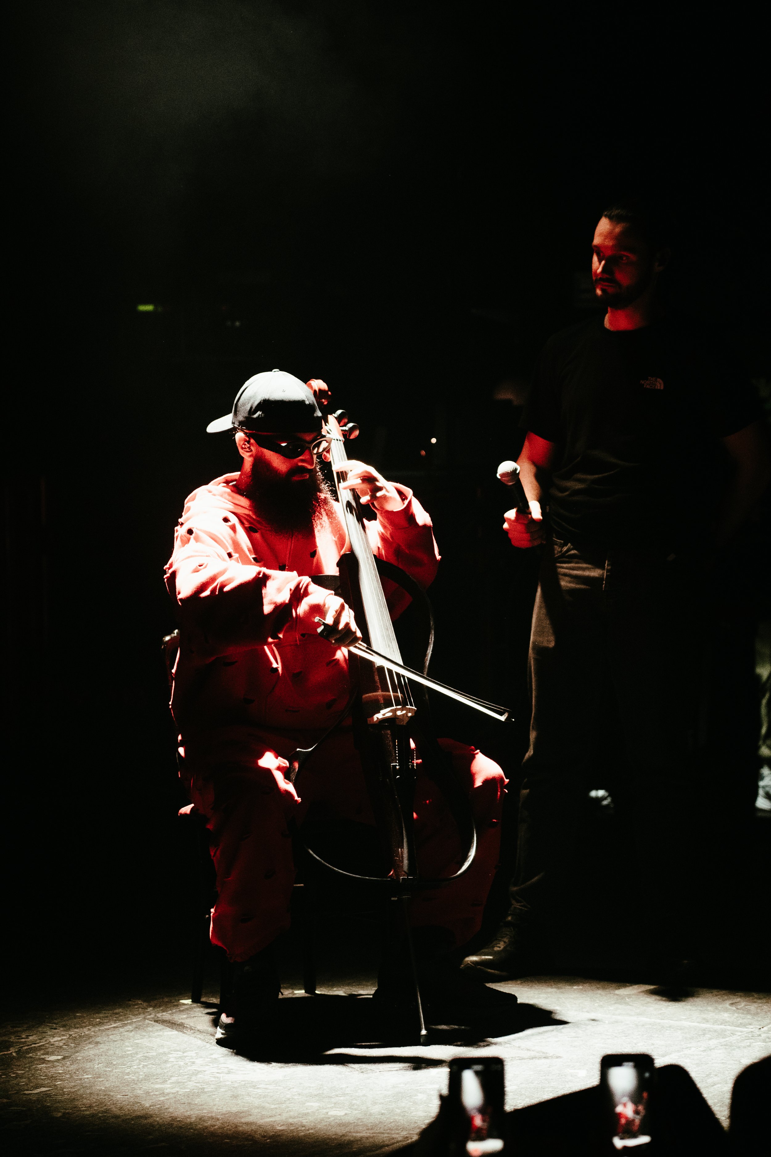 Un homme assis jouant du violoncelle sur scène, vêtu d'une capuche et d'une veste rouge, avec un autre homme debout à côté, tenant un microphone, dans une lumière tamisée.