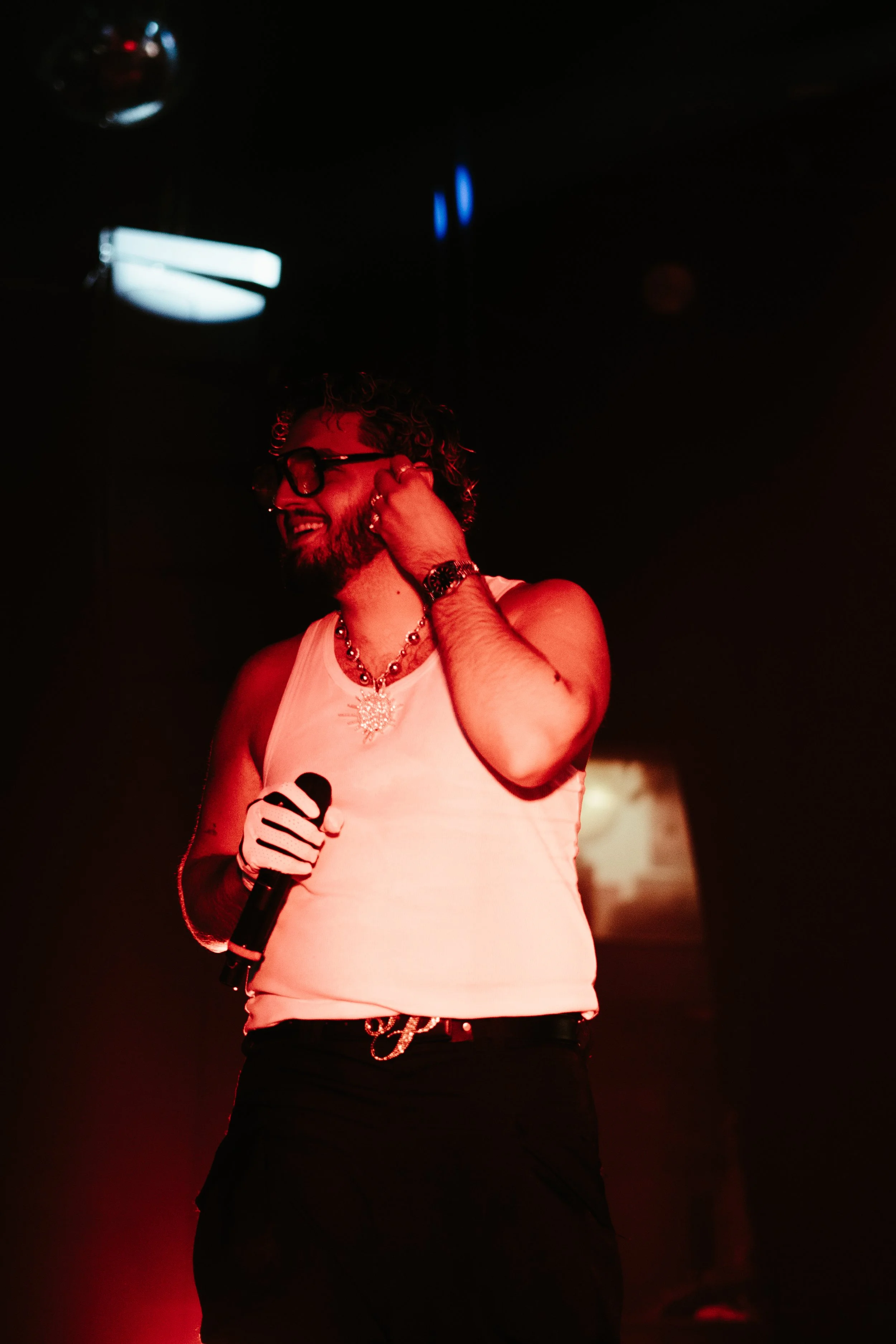 Un homme souriant avec des cheveux bouclés, portant des lunettes, un collier, une montre et un t-shirt blanc, tenant un micro, dans une ambiance lumineuse rouge dans une salle de spectacle ou un club.