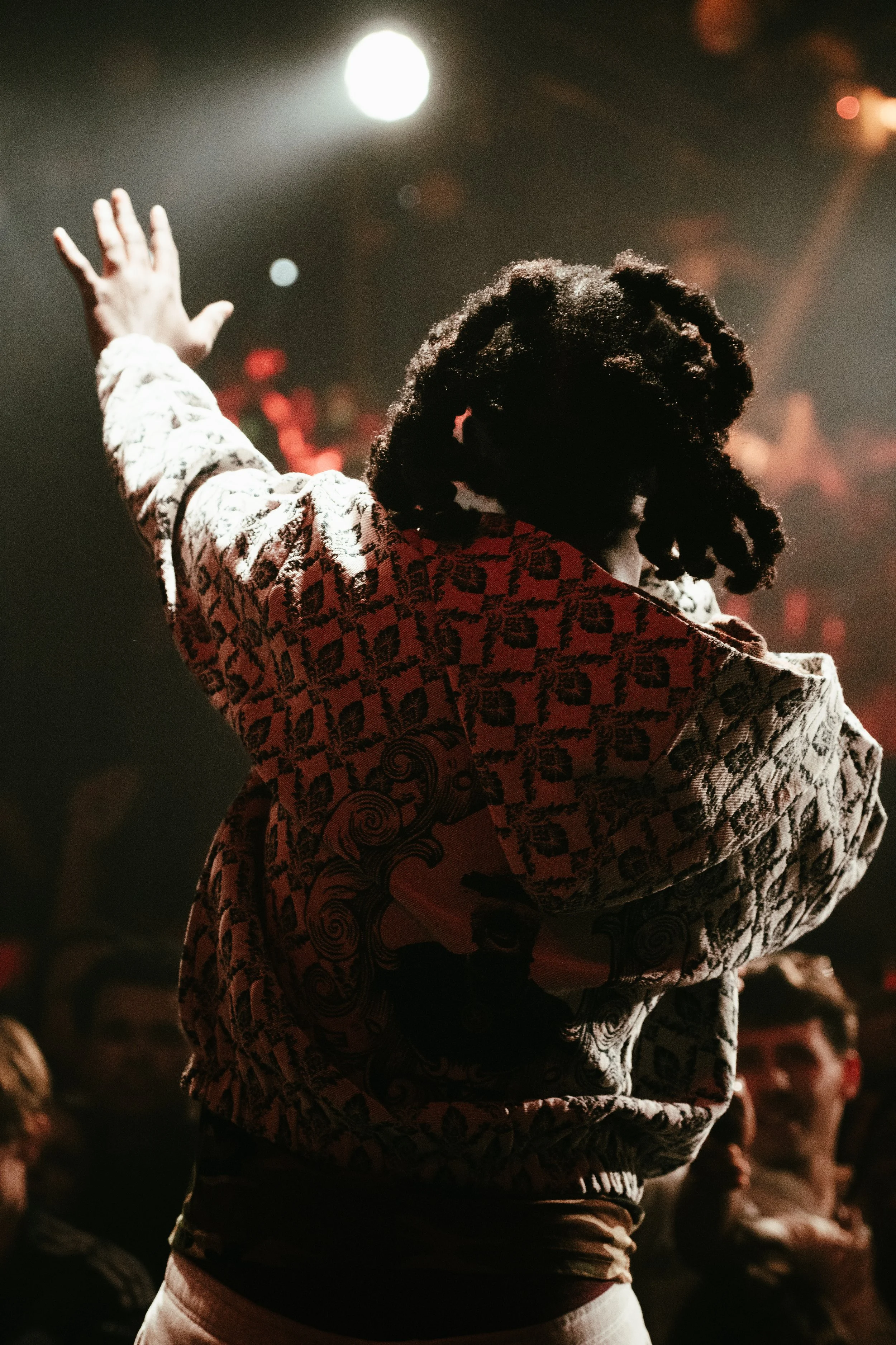 Une personne avec des cheveux bouclés, portant un sweat à capuche à motif, gesticulant dans une ambiance de spectacle ou de concert avec un éclairage tamisé.