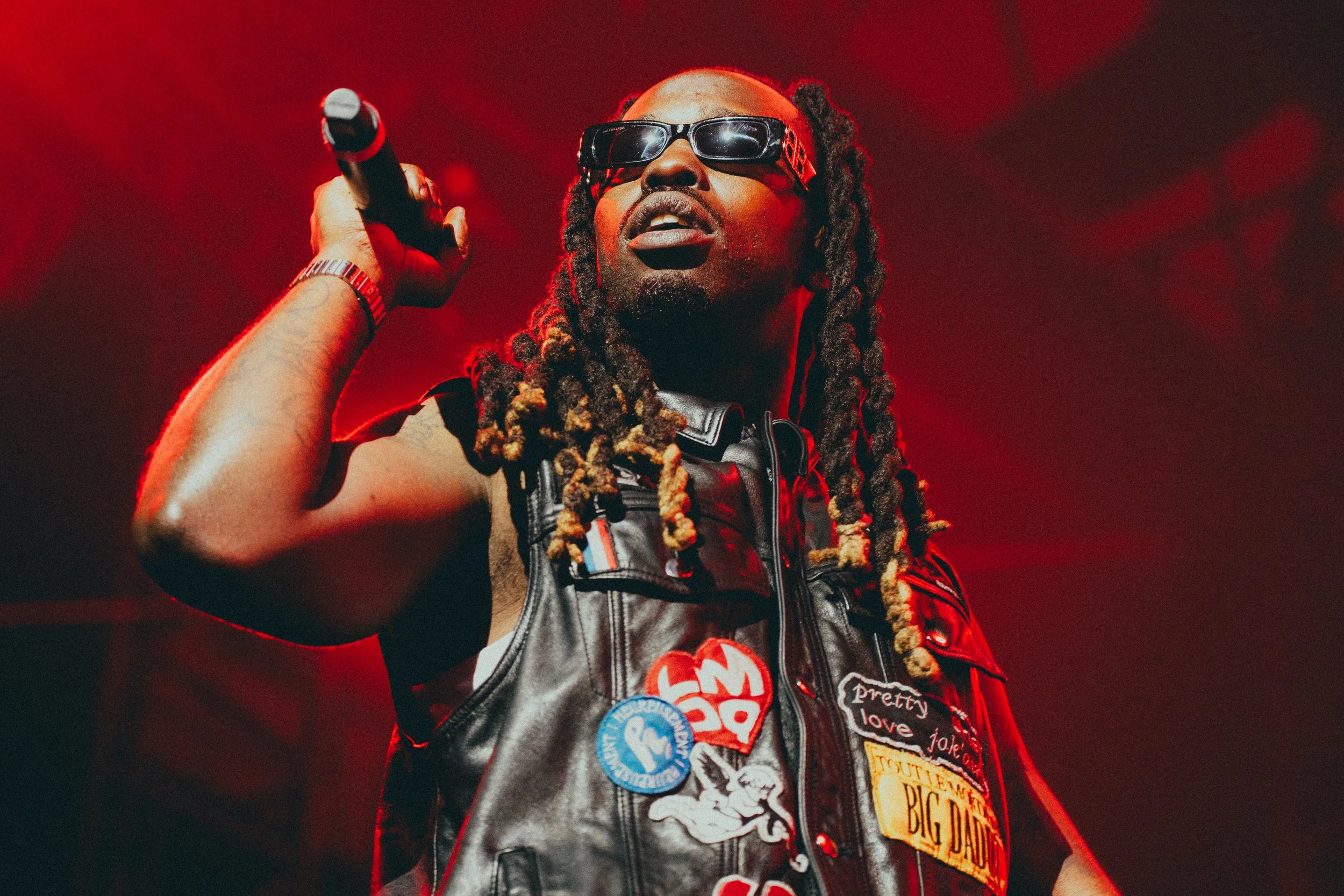 Un homme avec des dreadlocks portant des lunettes de soleil, une veste en cuir décorée de plusieurs badges, tenant un microphone dans sa main, sur scène avec un fond rouge.