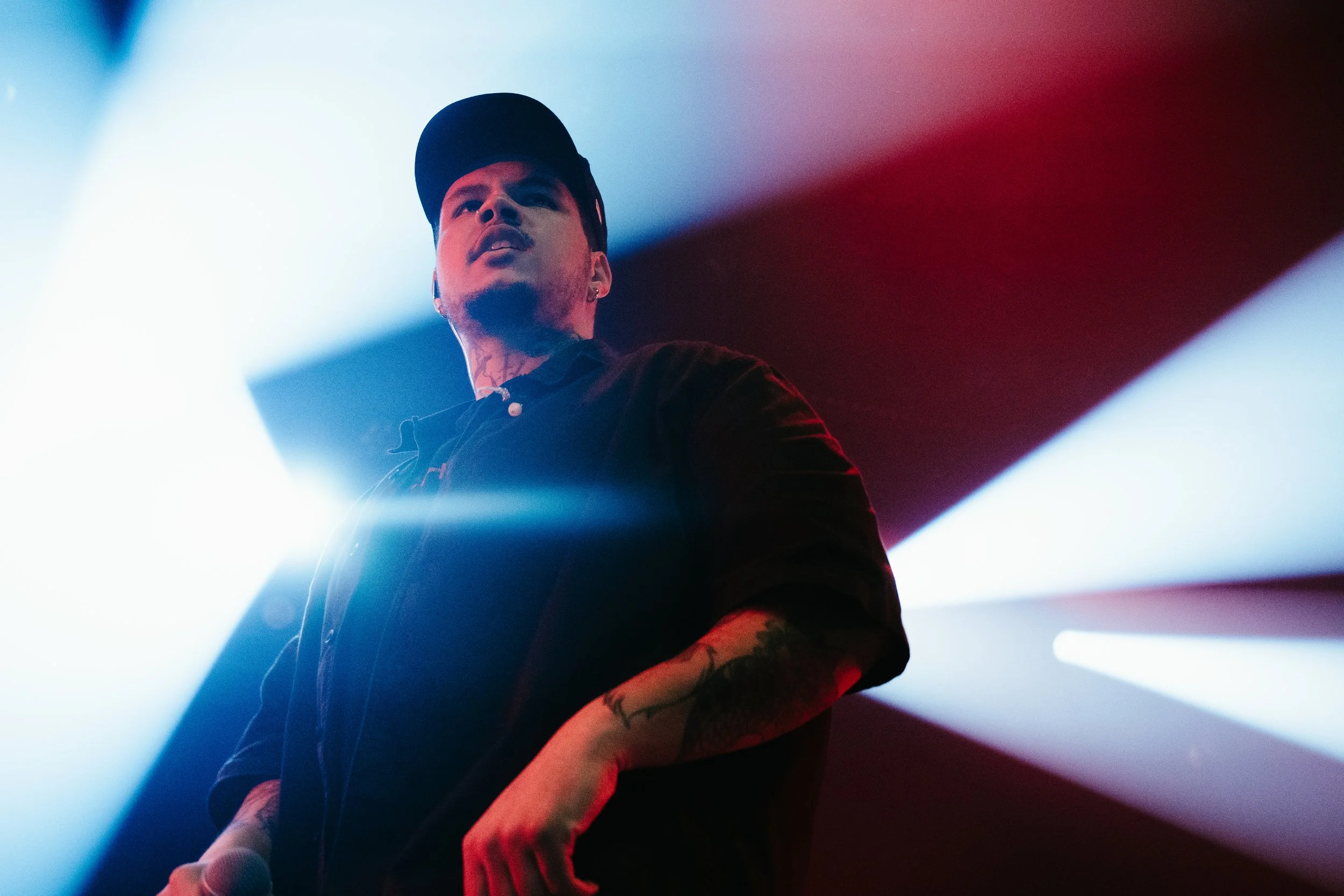 Un homme portant une casquette noire, avec des tatouages visibles sur le bras, est sur scène sous des lumières de couleurs rouge, blanc et bleu.