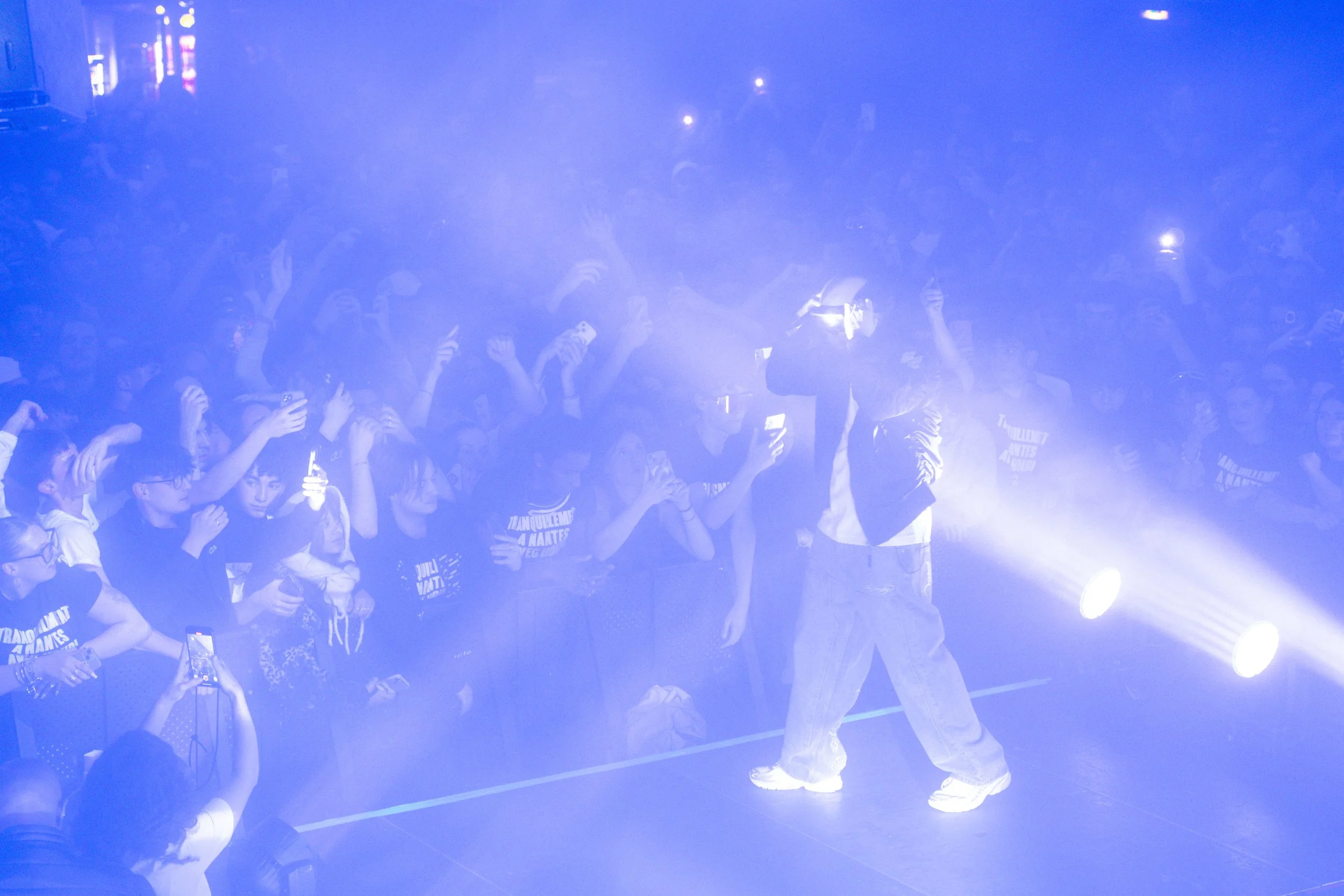 Un artiste en sweat, casquette et jean large performe sur scène avec des lumières brillantes, devant une foule de spectateurs en T-shirts noirs, certains prennent des photos ou vidéos avec leur téléphone.