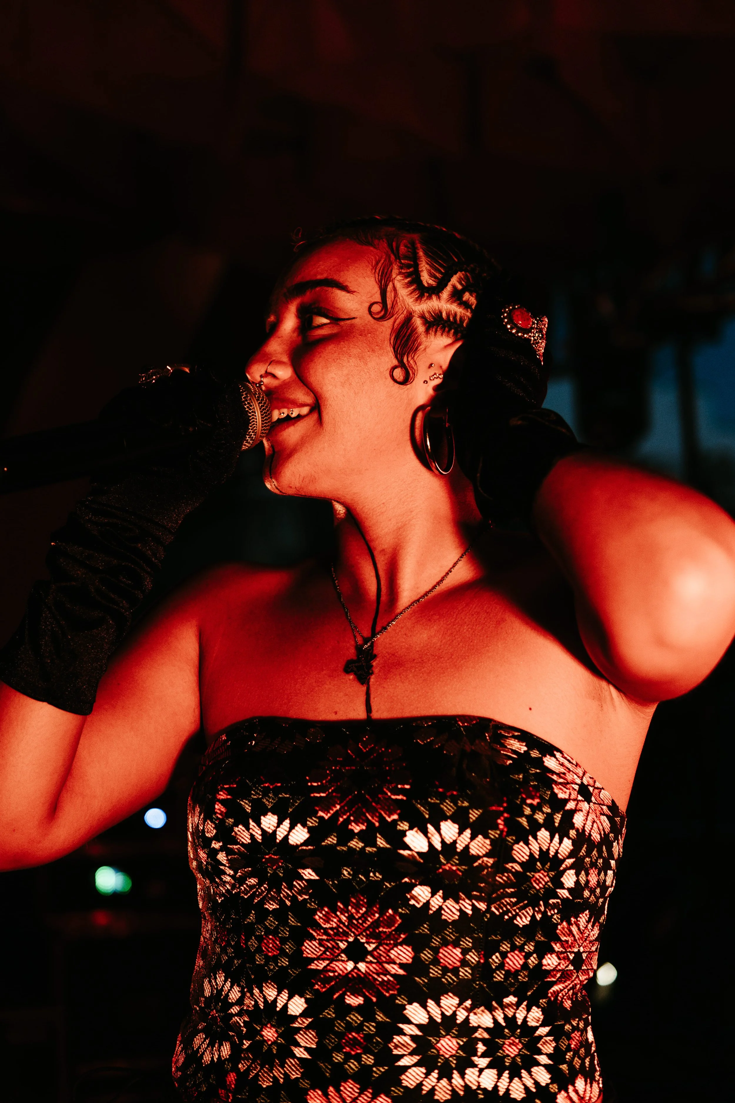 Jeune femme chantant dans un micro lors d'une performance, portant un vêtement moulant à motif géométrique, portant des boucles d'oreilles et des bijoux, avec des cheveux peignés en motifs.