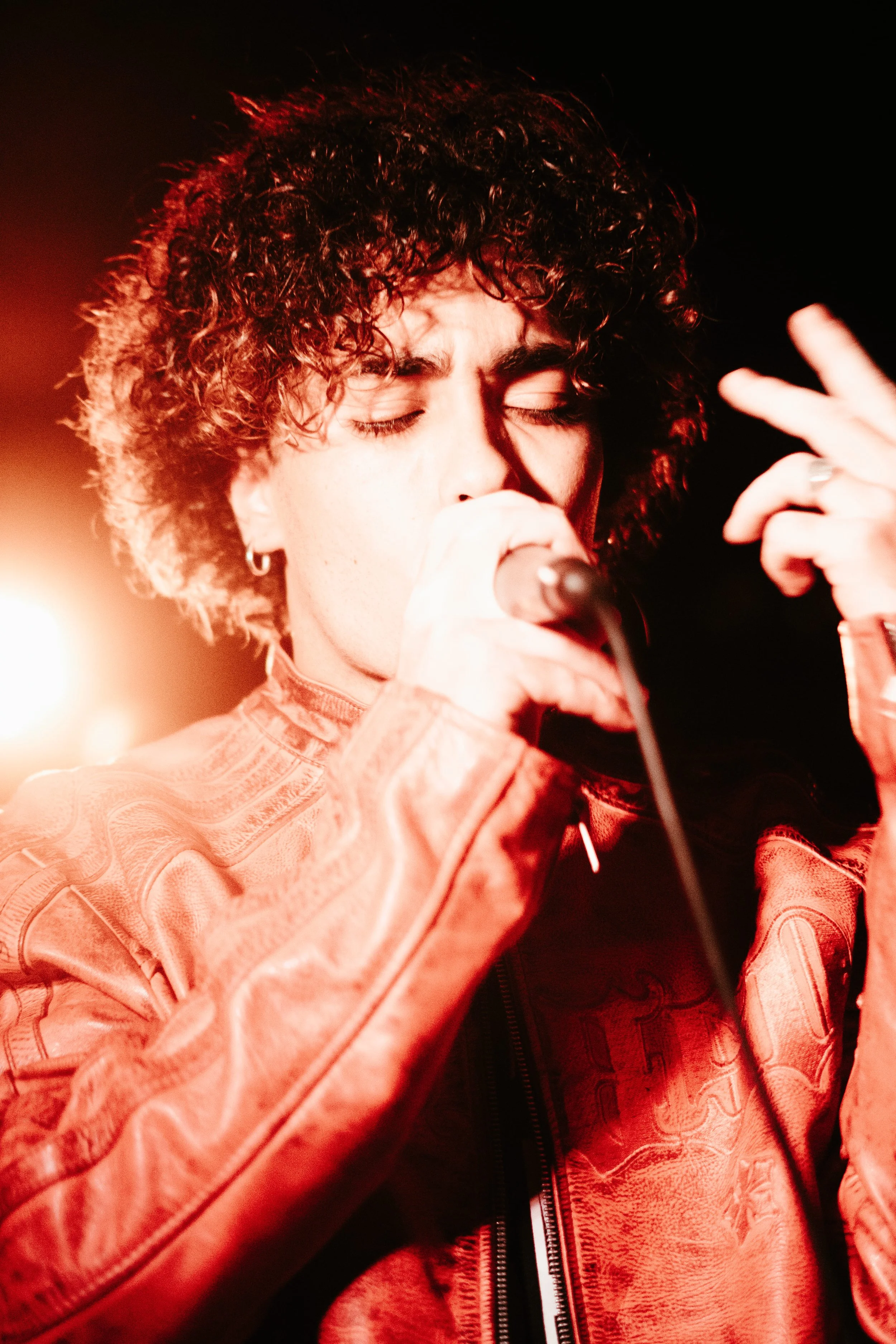 Un jeune homme avec des cheveux bouclés chante dans un microphone avec ses yeux fermés, portant une veste en cuir rouge et faisant un geste avec sa main droite.