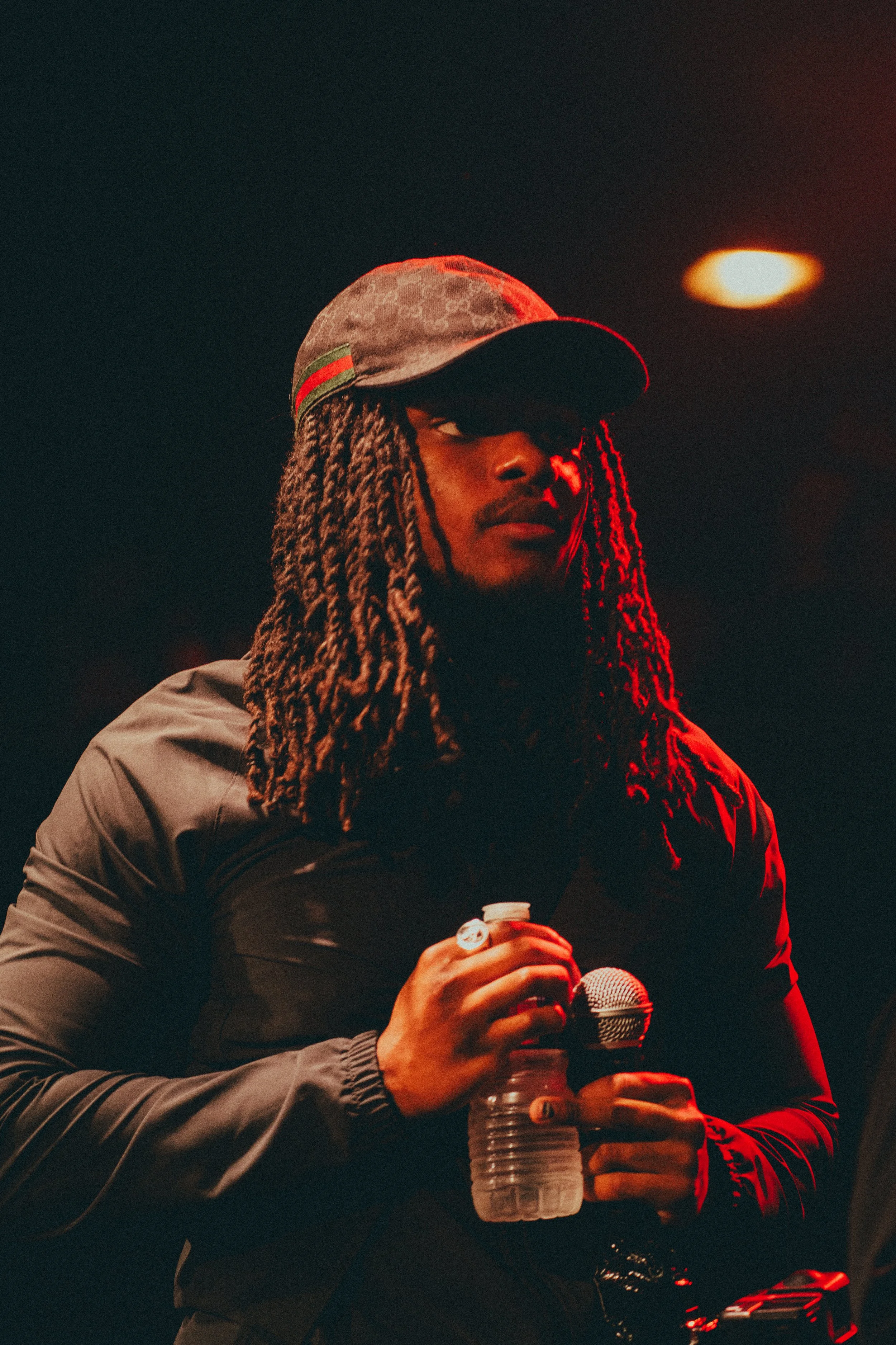 Jeune homme portant une casquette, tenant une bouteille d'eau et un microphone, dans un environnement sombre avec un éclairage rouge.