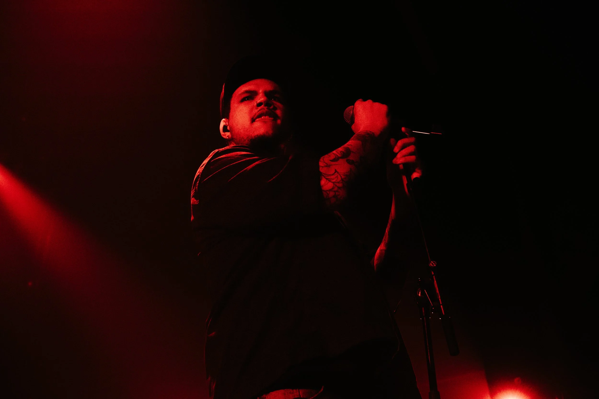 Homme chantant dans une scène sombre éclairée en rouge, tenant un microphone avec des tatouages visibles sur le bras.