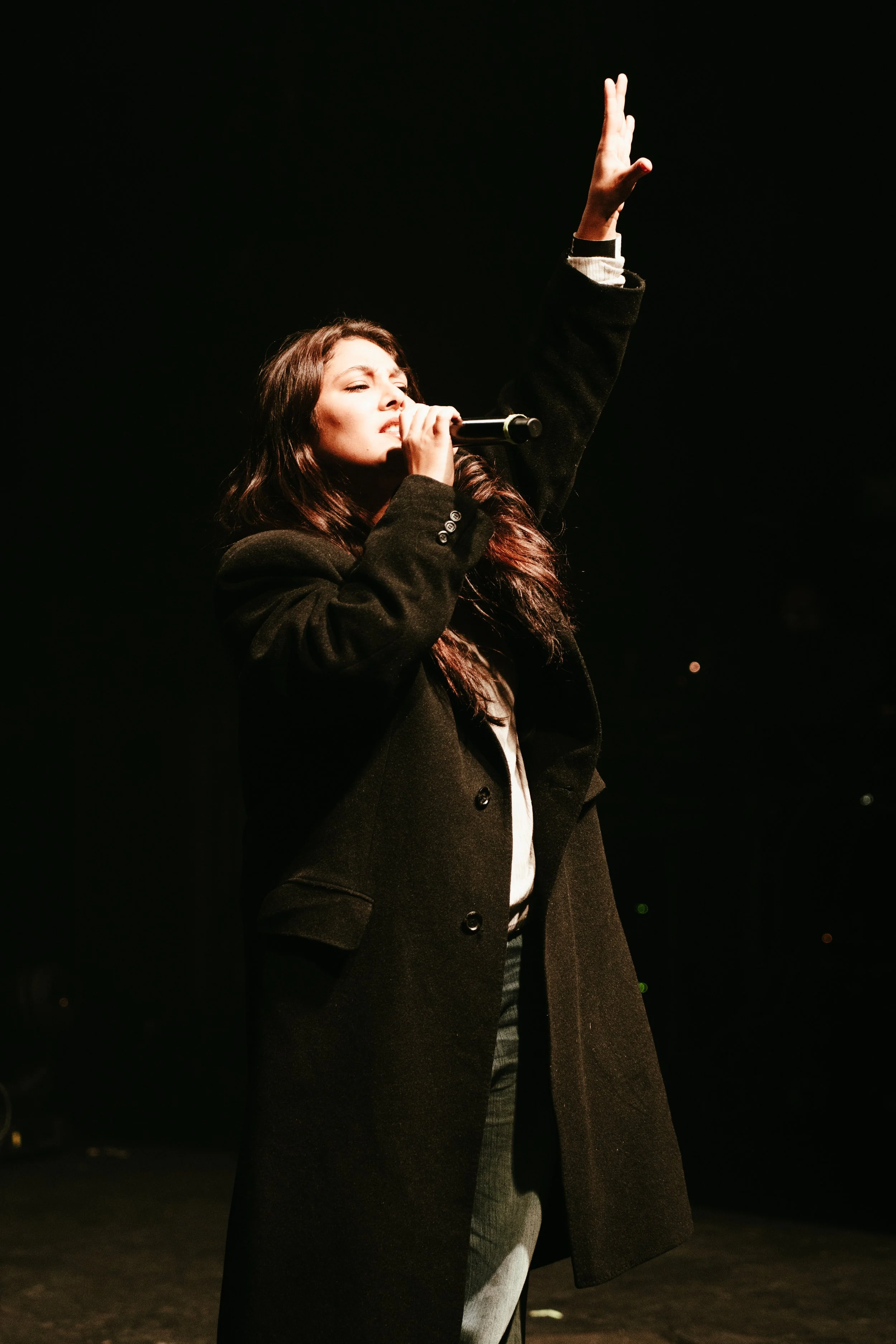 Une femme chante avec un microphone sur scène, portant un manteau noir et levant la main.
