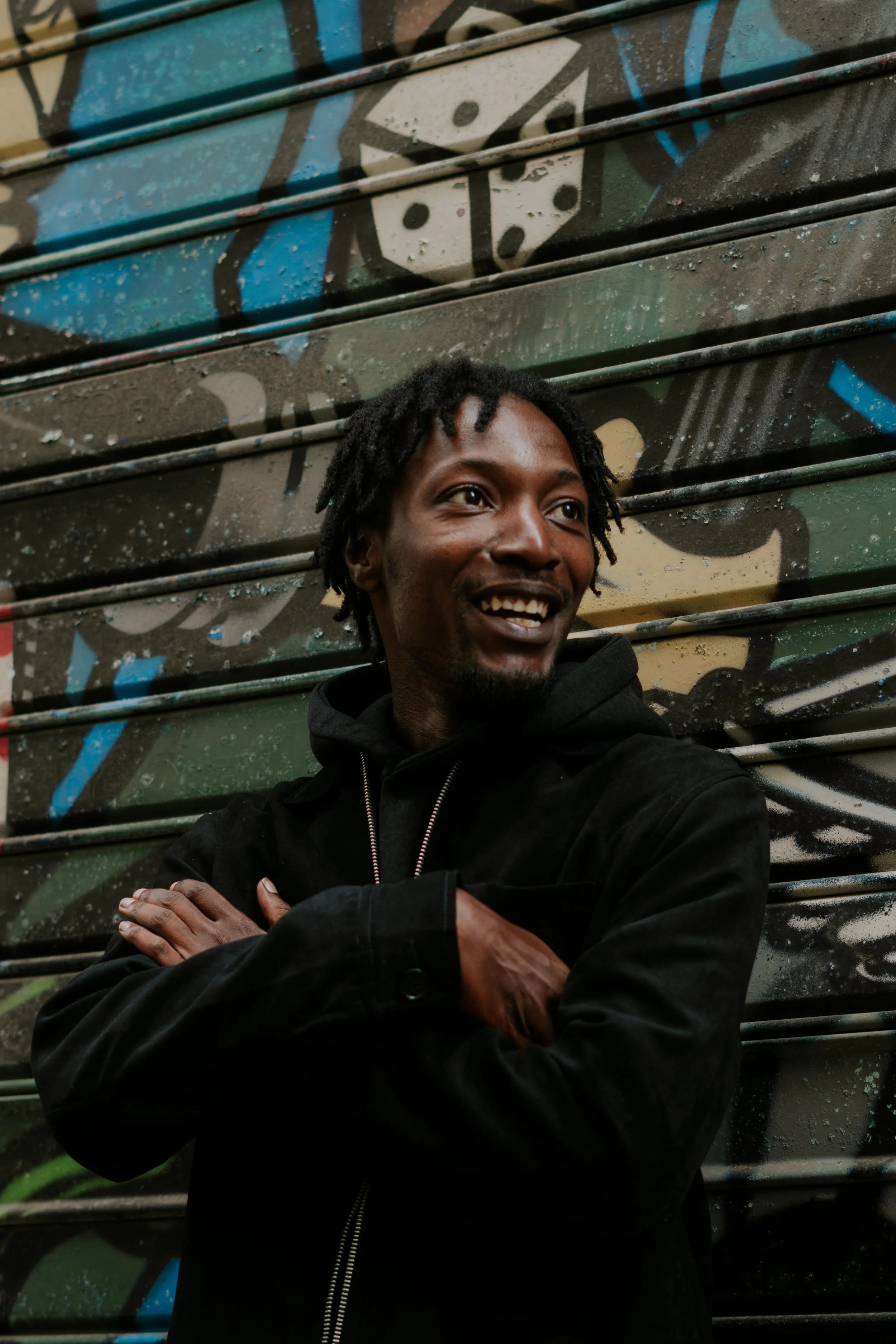 Un homme souriant avec des dreadlocks, portant une veste noire, se tient devant un mur avec un graffiti coloré.