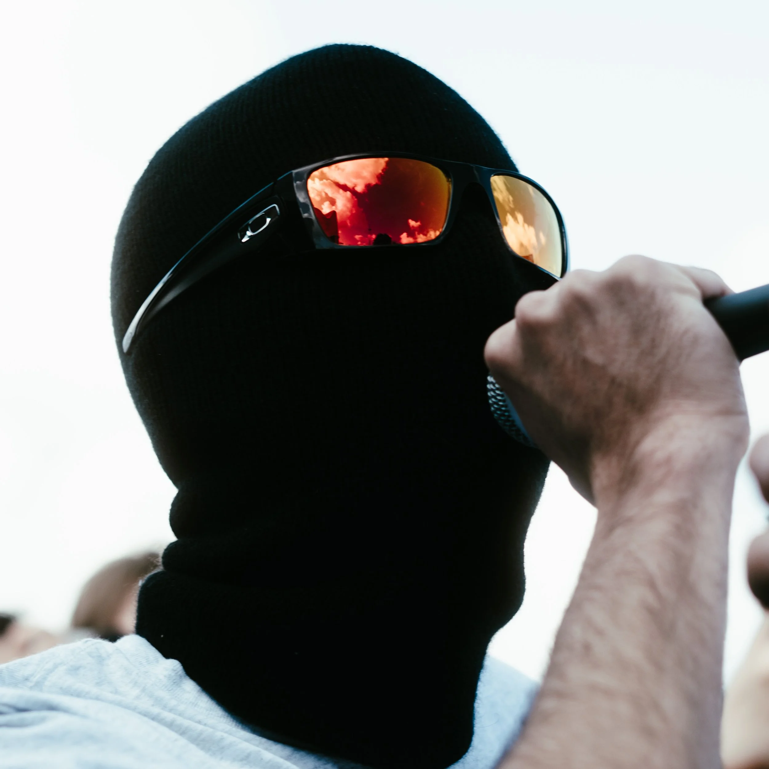 Personne portant un masque noir, lunettes de soleil avec reflet orange, tenant un microphone.