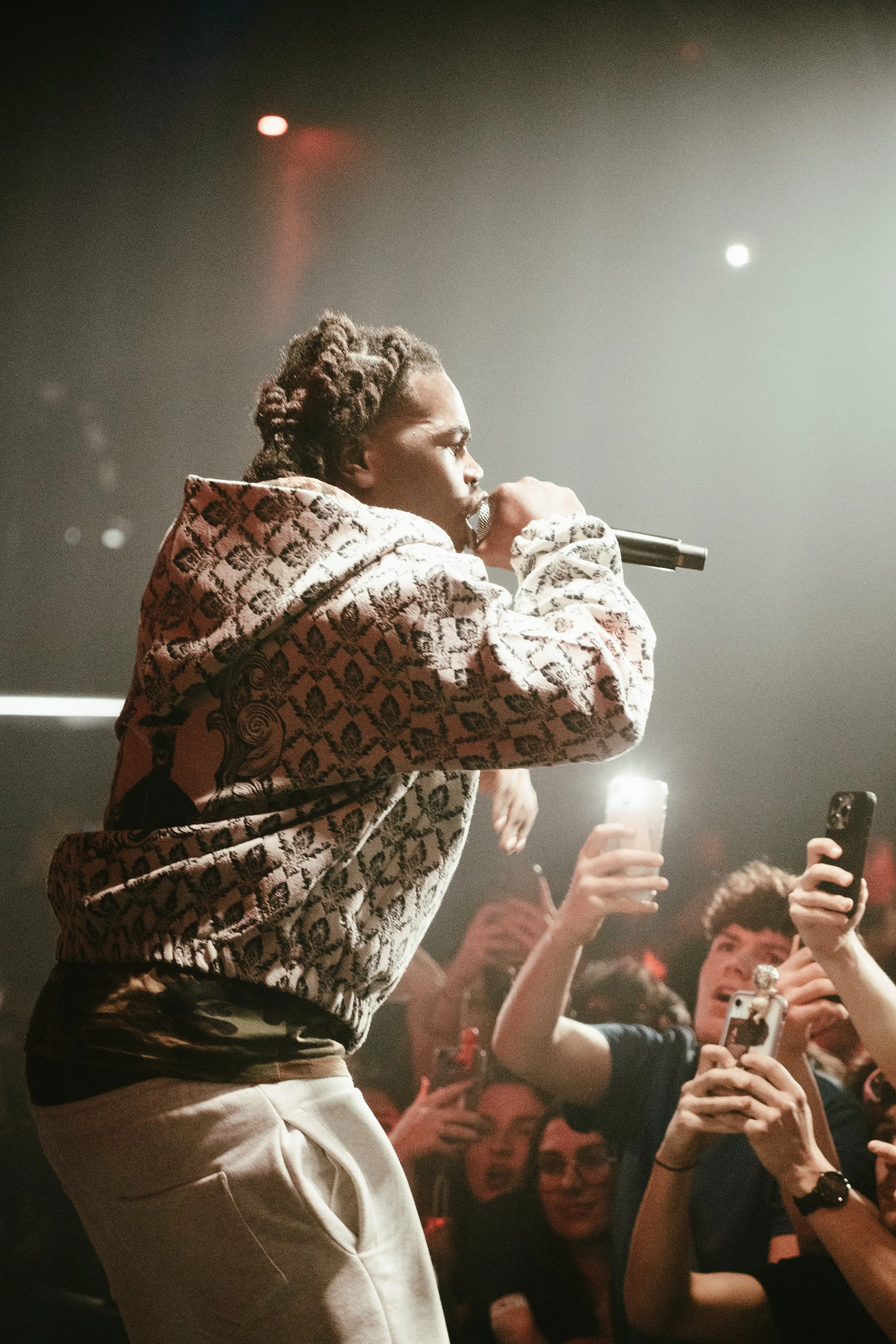 Un artiste de hip-hop avec des dreadlocks, portant un sweat à motif, qui chante avec un microphone sur scène, entouré de fans prenant des photos et vidéos avec leurs téléphones lors d’un concert.