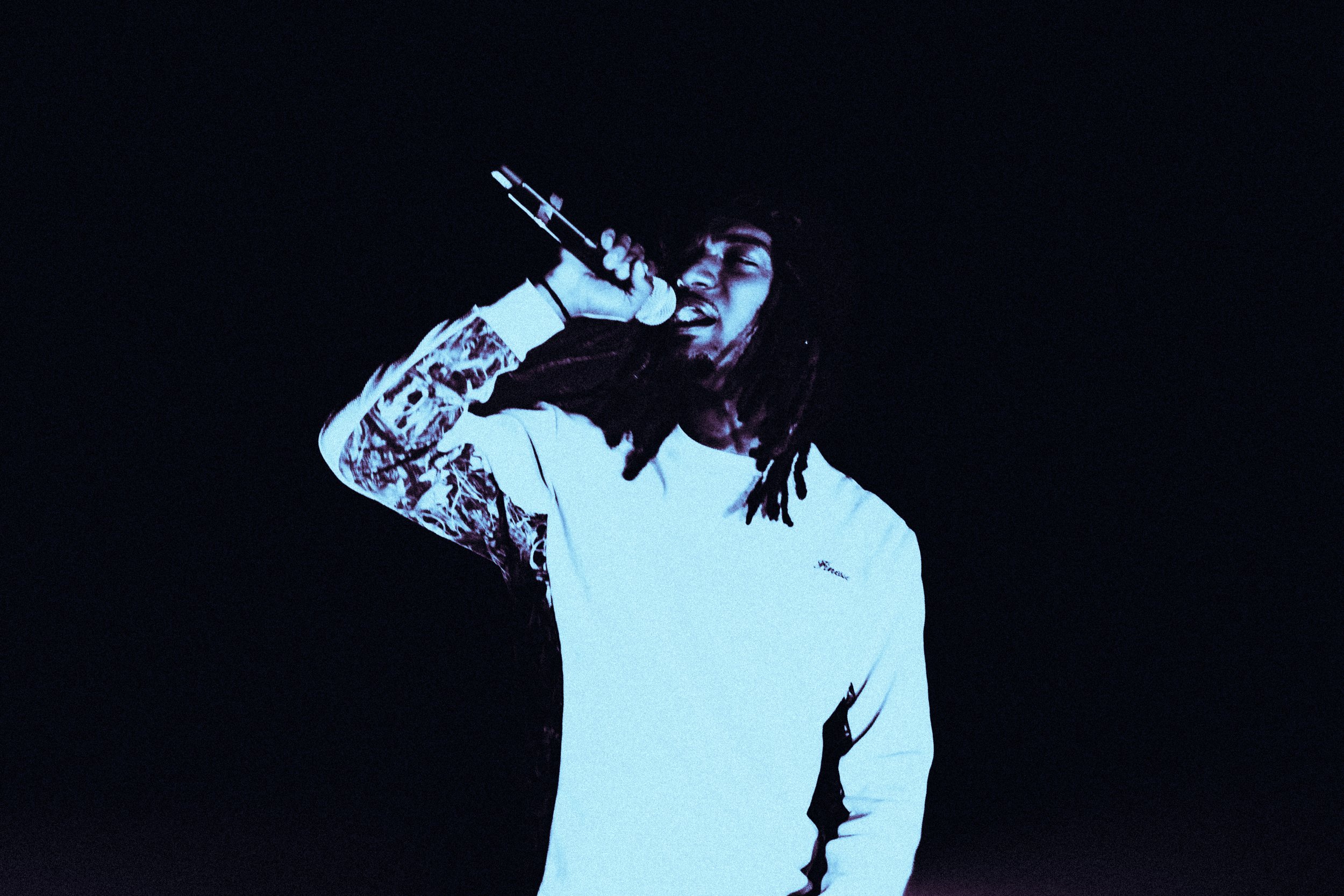 Un homme à dreadlocks chante dans un micro contre un fond noir.