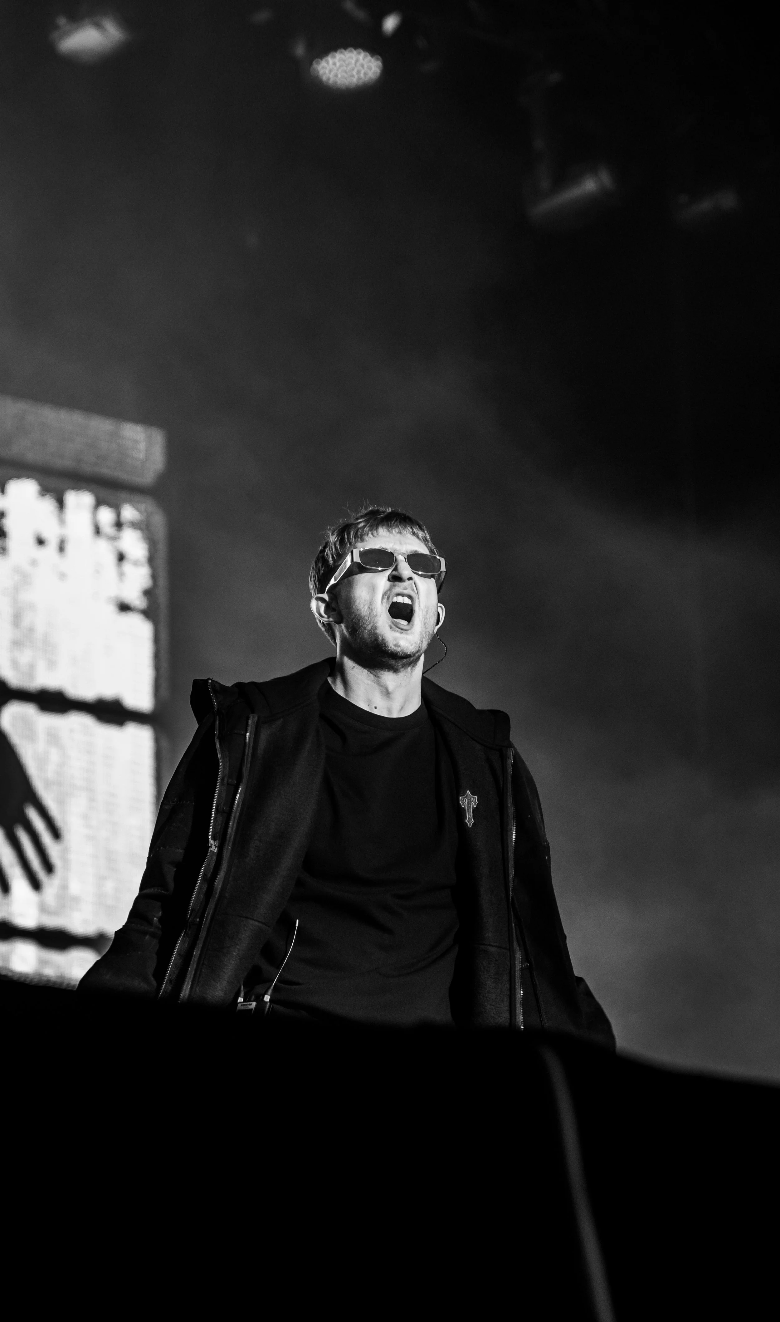 Un homme portant des lunettes de soleil chante ou crie lors d'une performance en noir et blanc dans une ambiance de concert avec un fond lumineux.