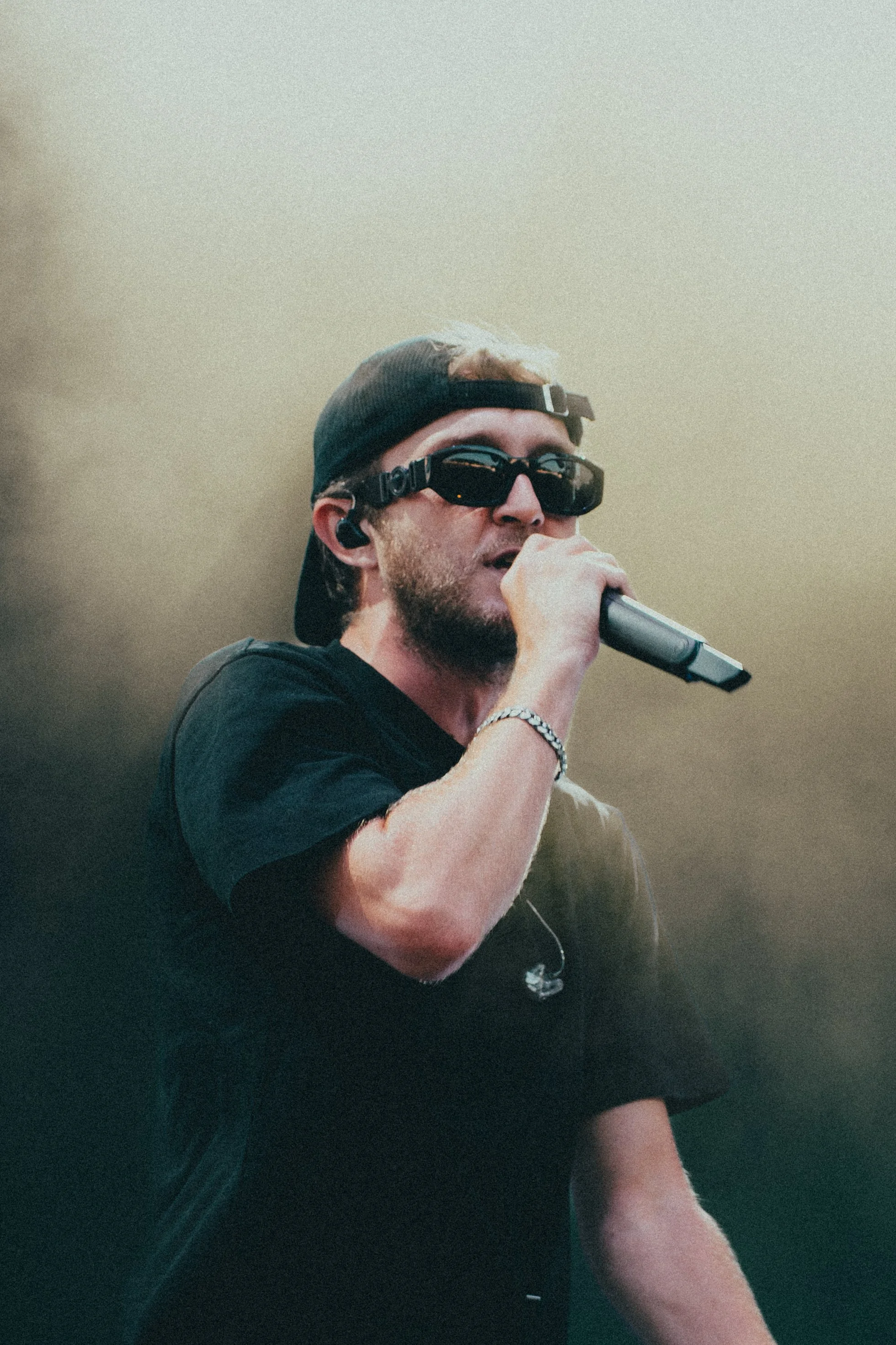 Un homme portant un casque noir, des lunettes de soleil noires, une t-shirt noir, un bracelet en argent, et une boucle d'oreille, chantant ou parlant dans un microphone lors d'une performance ou d'un événement.