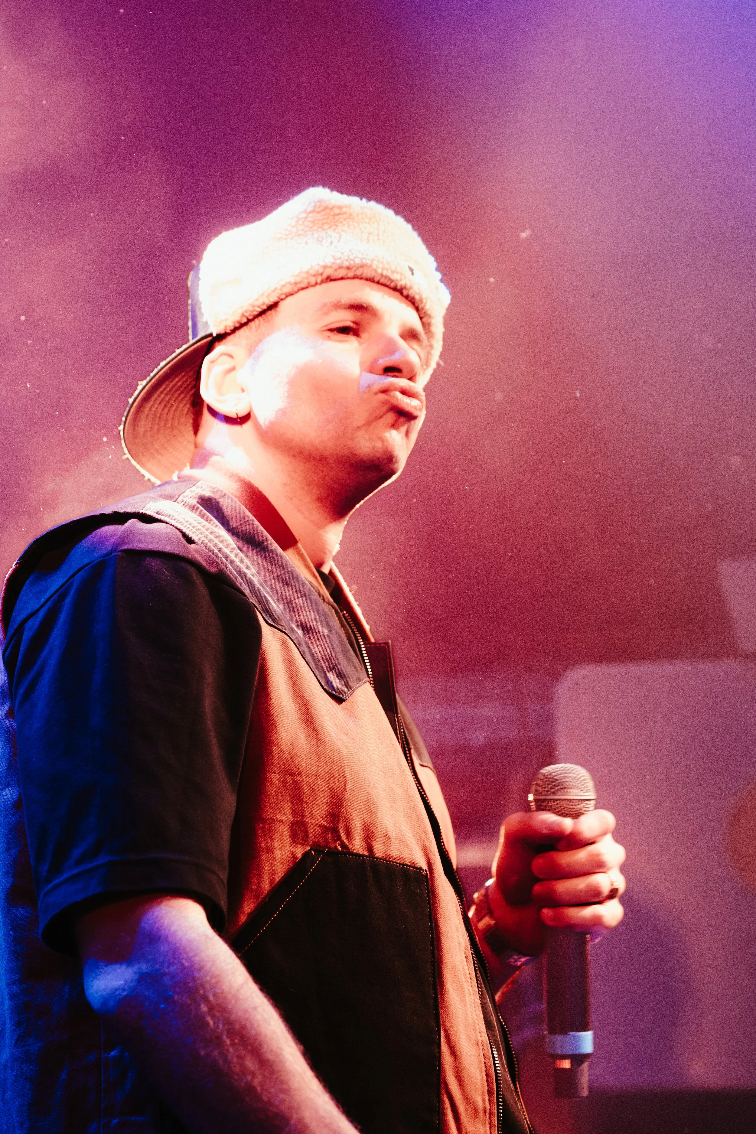 Un homme tenant un microphone, portant un pull à manches courtes et une casquette, lors d'une performance sur scène avec un éclairage rose et violet.
