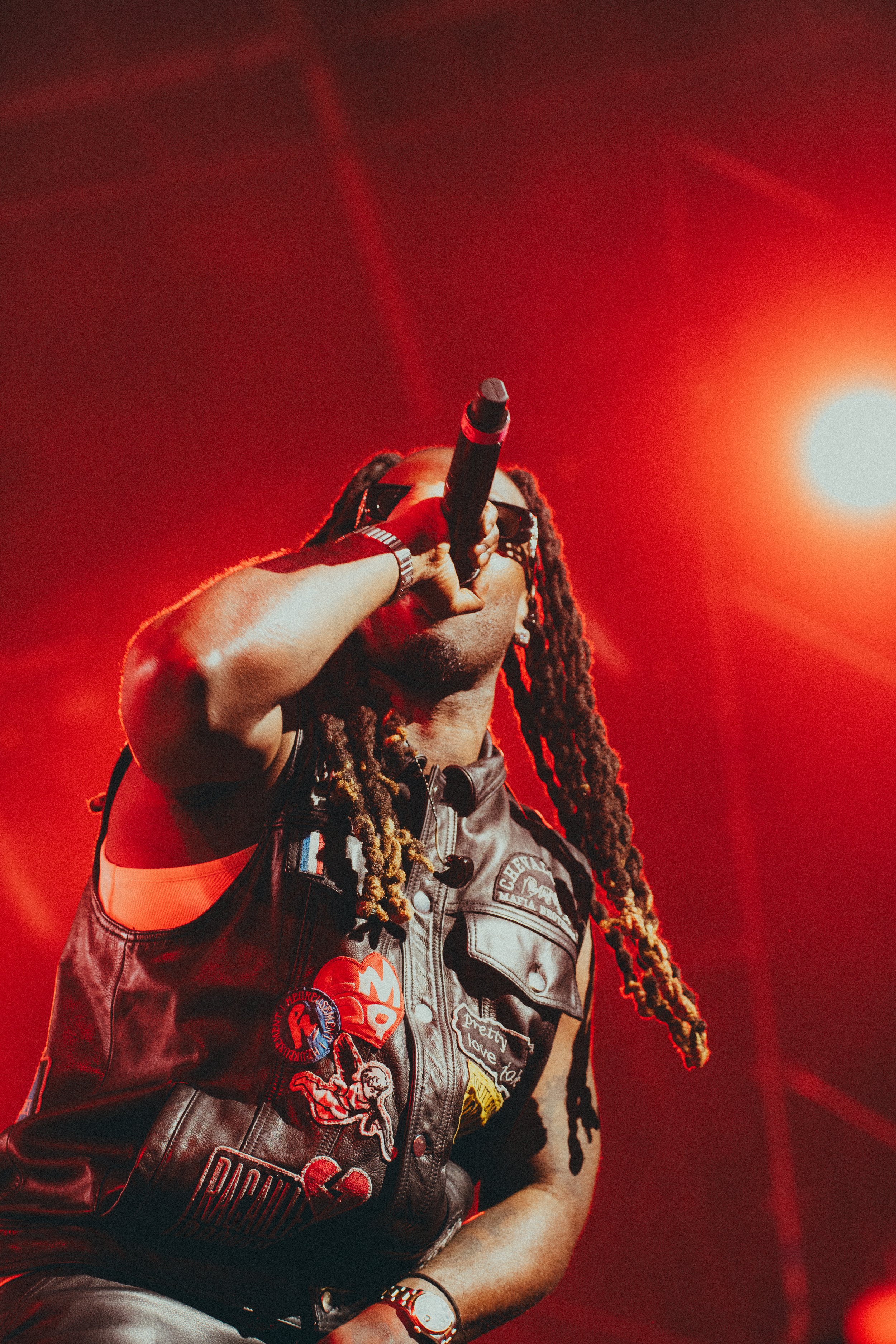 Un chanteur avec de longs dreadlocks, portant une veste en cuir décorée de broderies et de badges, chante dans un micro sous un éclairage rouge vif.