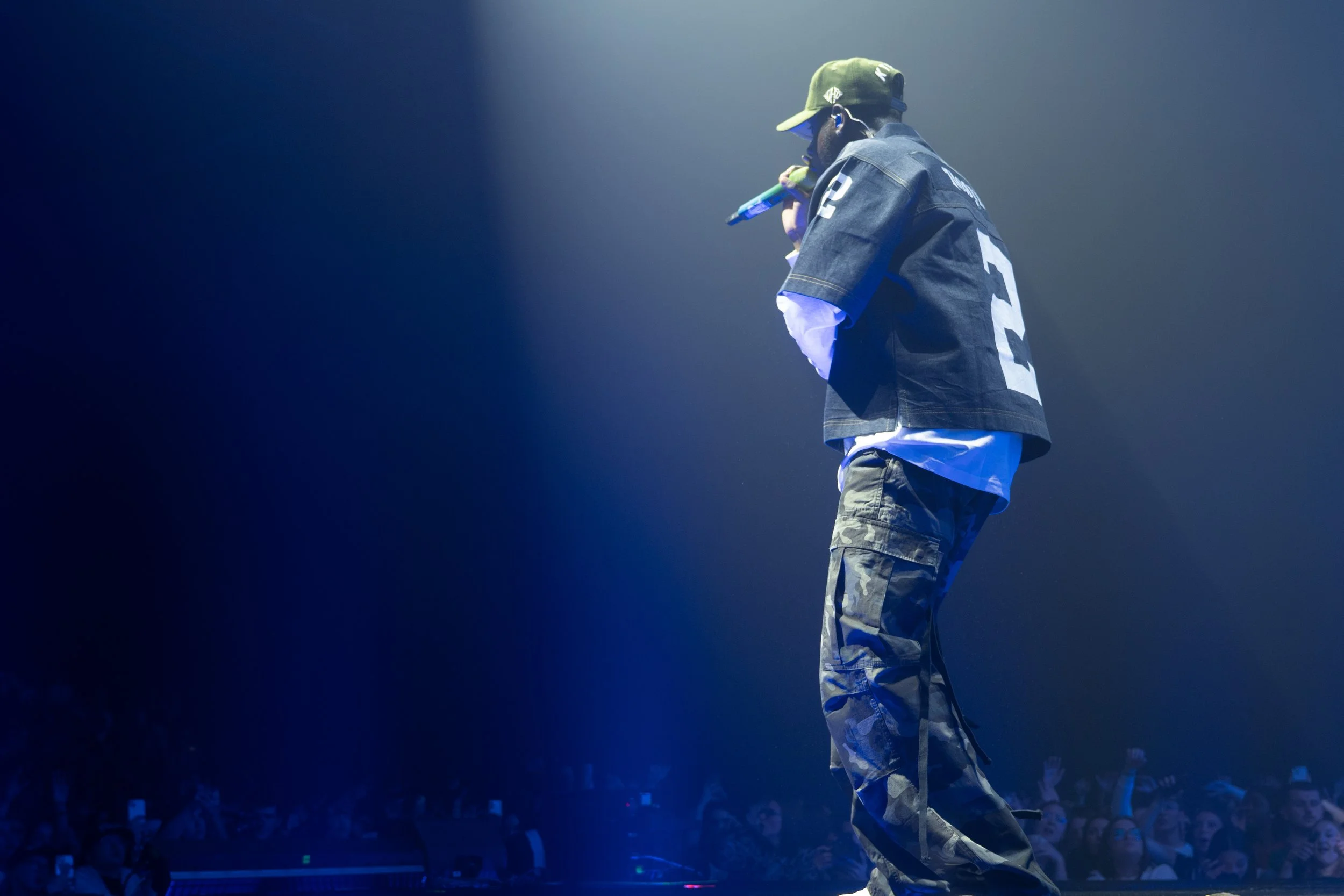 Un artiste sur scène tenant un microphone dans une main, portant une casquette, un pantalon camouflage, un sweatshirt avec le chiffre 2, et un veston noir, dans une lumière tamisée avec un public en arrière-plan.