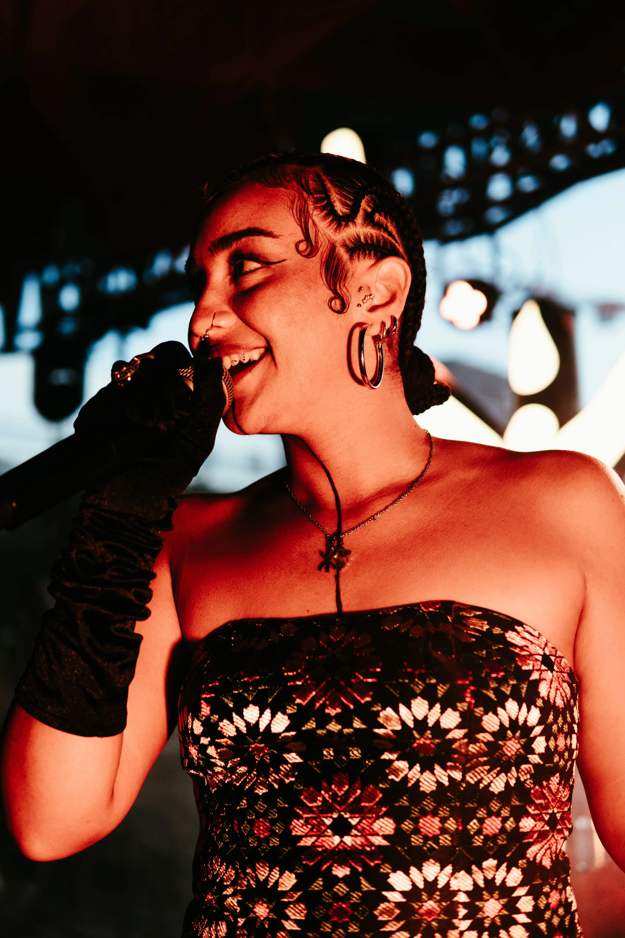 Jeune femme avec des tatouages, plusieurs piercings, coiffure en braids et boucle d'oreilles, en train de chanter ou parler en microphone dans un environnement lumineux