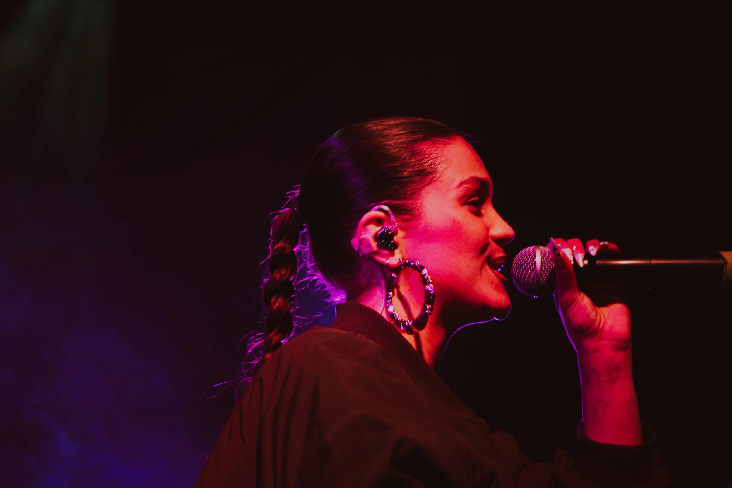 Une femme chante en microphone, portant de grosses boucles d'oreilles, avec un éclairage rose et violet dans un environnement sombre.