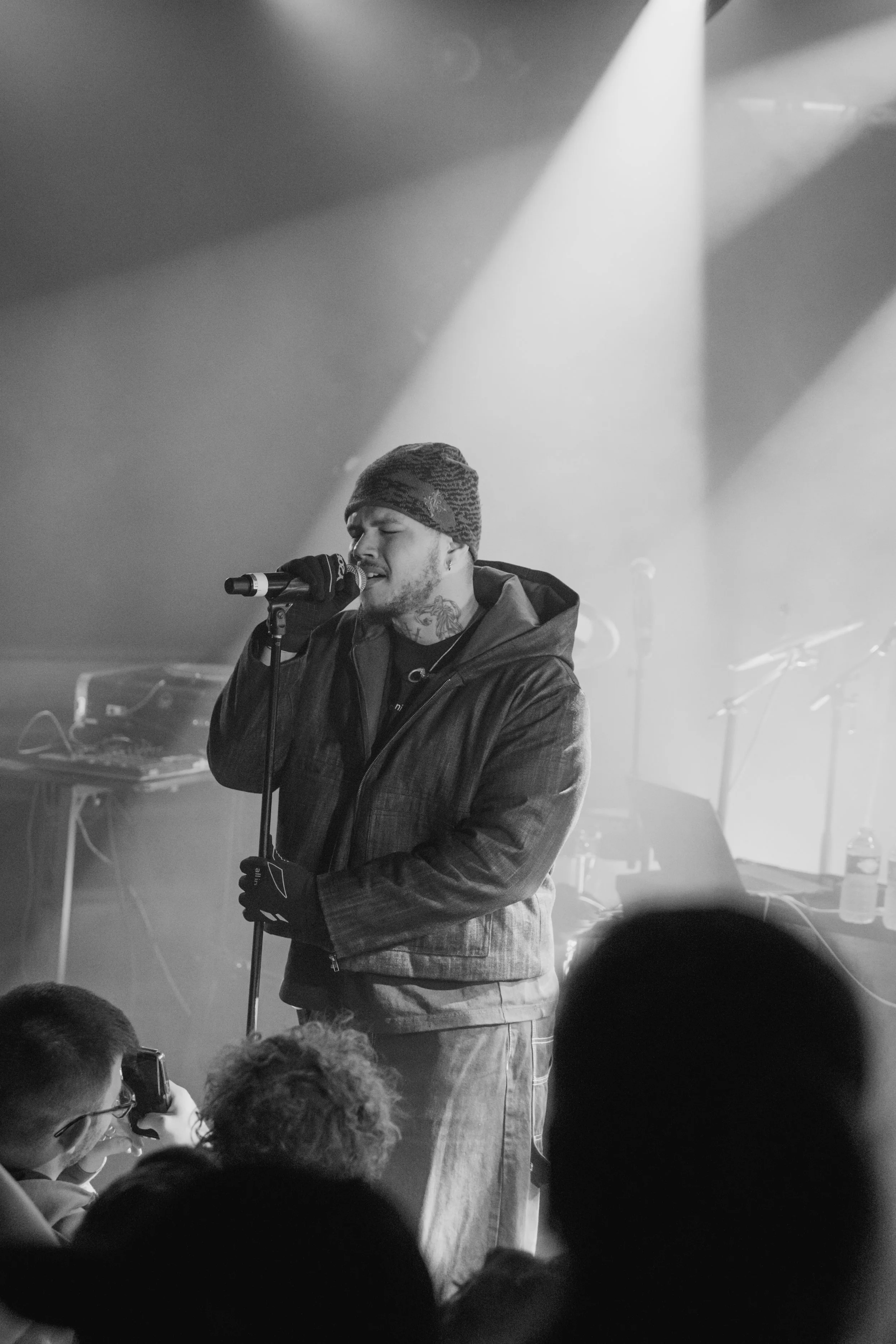 Un chanteur masculin en noir et blanc, portant un bonnet en laine, un manteau et des gants, chantant dans un micro devant un public dans une salle de concert