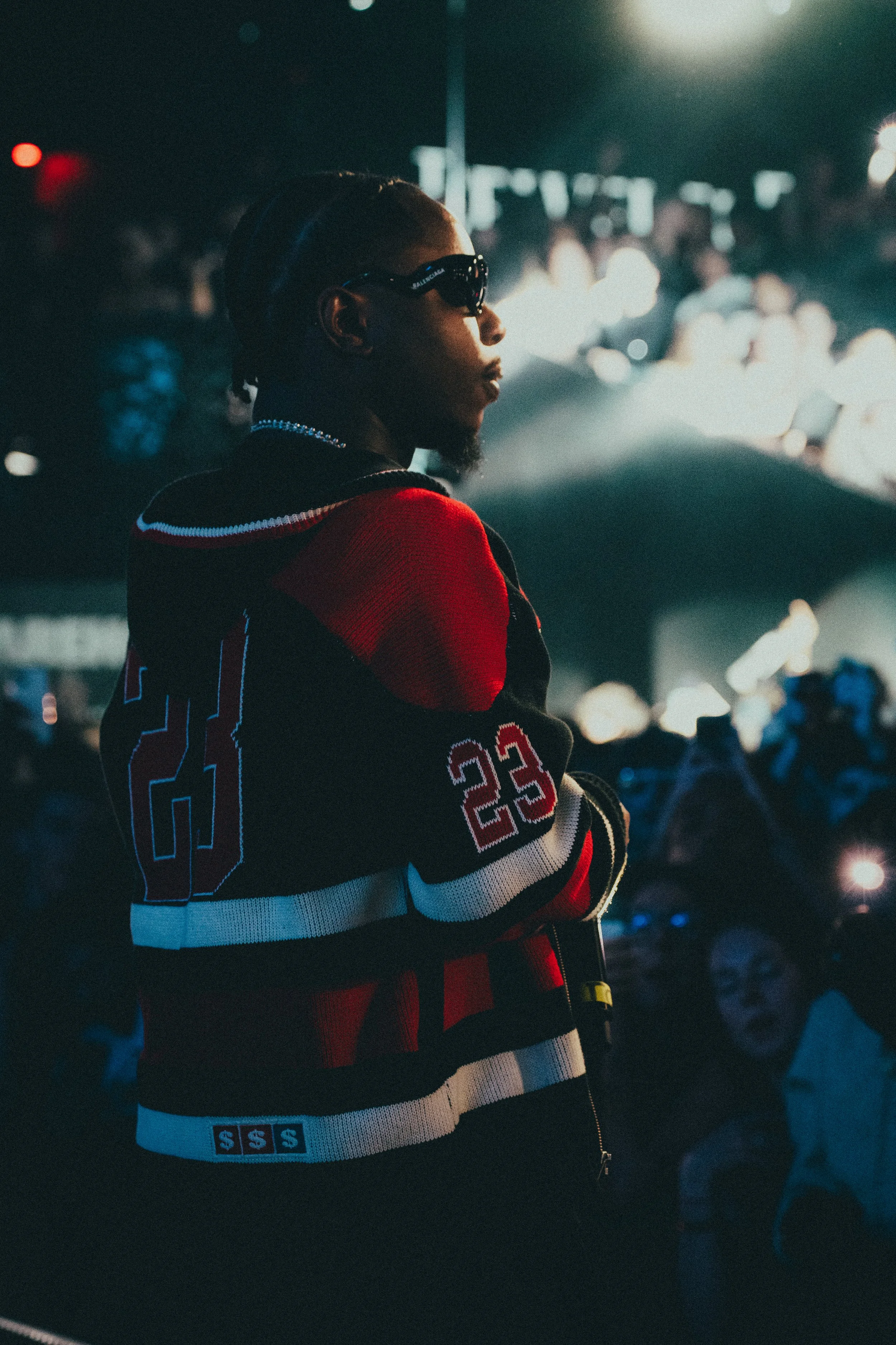 Un homme portant des lunettes de soleil et un sweat à capuche coloré, posé lors d'un événement nocturne avec une foule en arrière-plan.