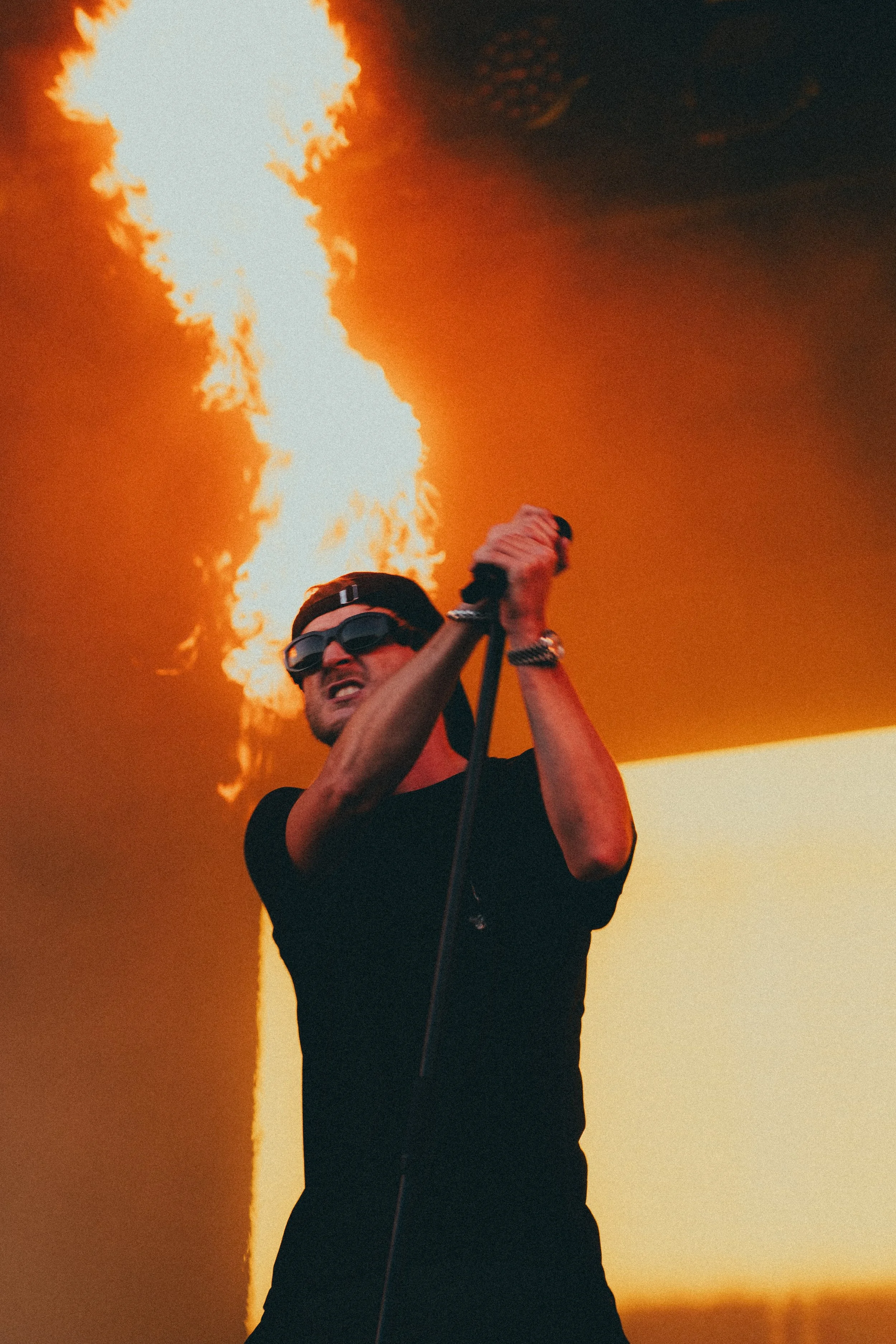 Un homme portant des lunettes noires et un t-shirt noir chante en tenant un micro avec un feu en arrière-plan.