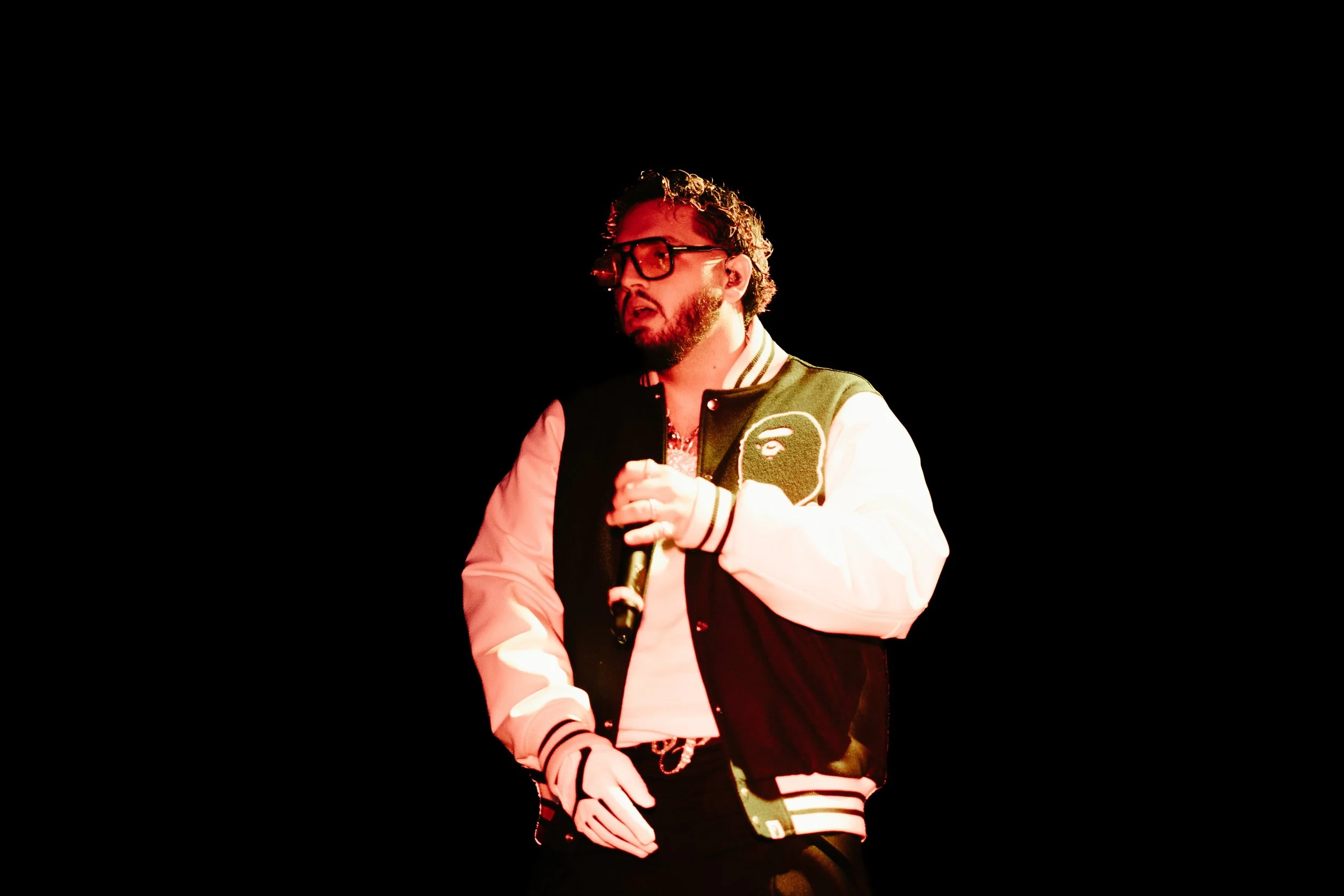 Un homme portant des lunettes, une veste à manches longues avec une tête de gorille brodée sur le devant, tient un microphone devant un mur noir, en train de chanter ou de parler.