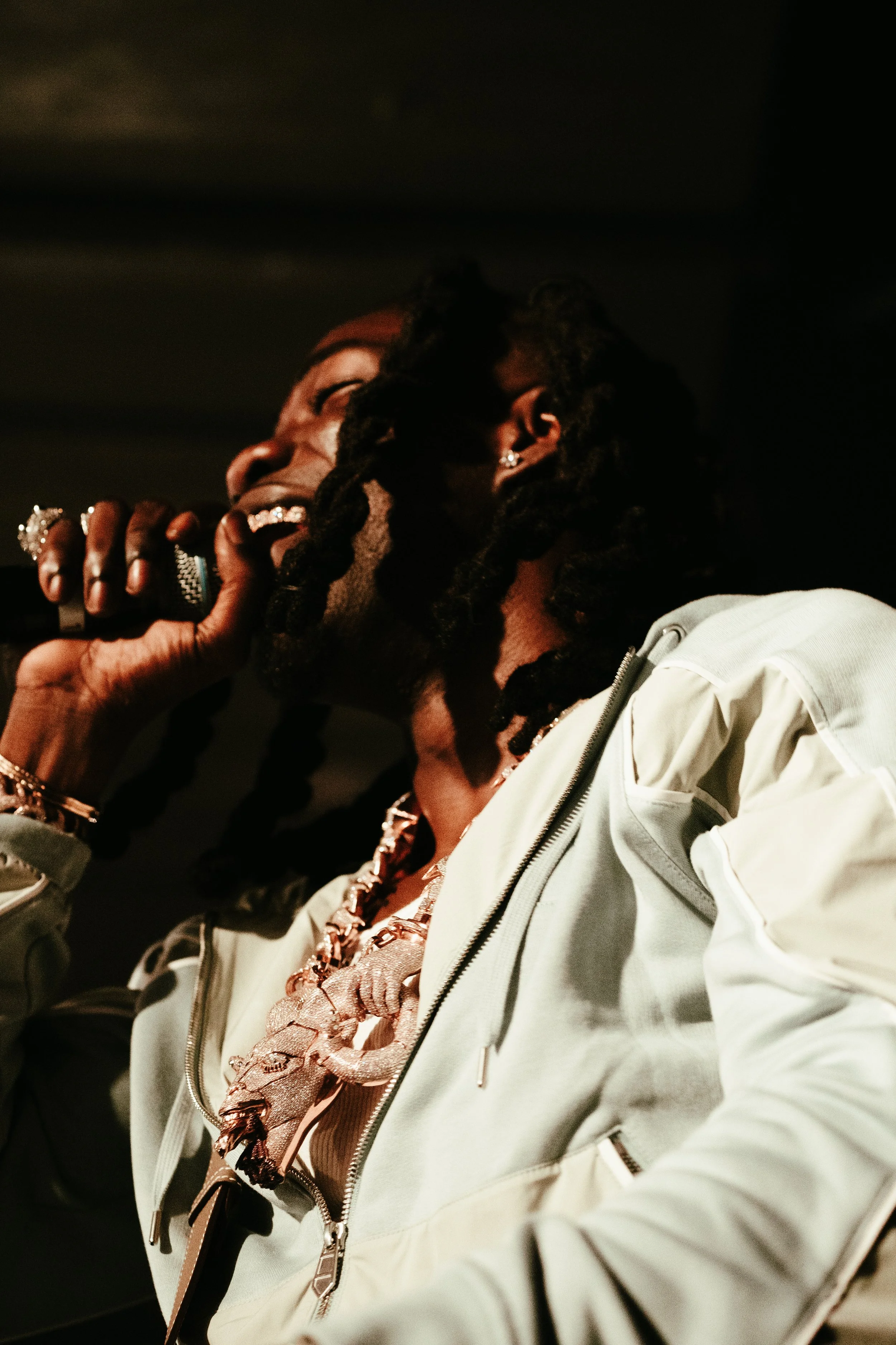 Une femme avec des cheveux en dreadlocks chante en tenant un microphone, portant une chaîne en or et une veste blanche.