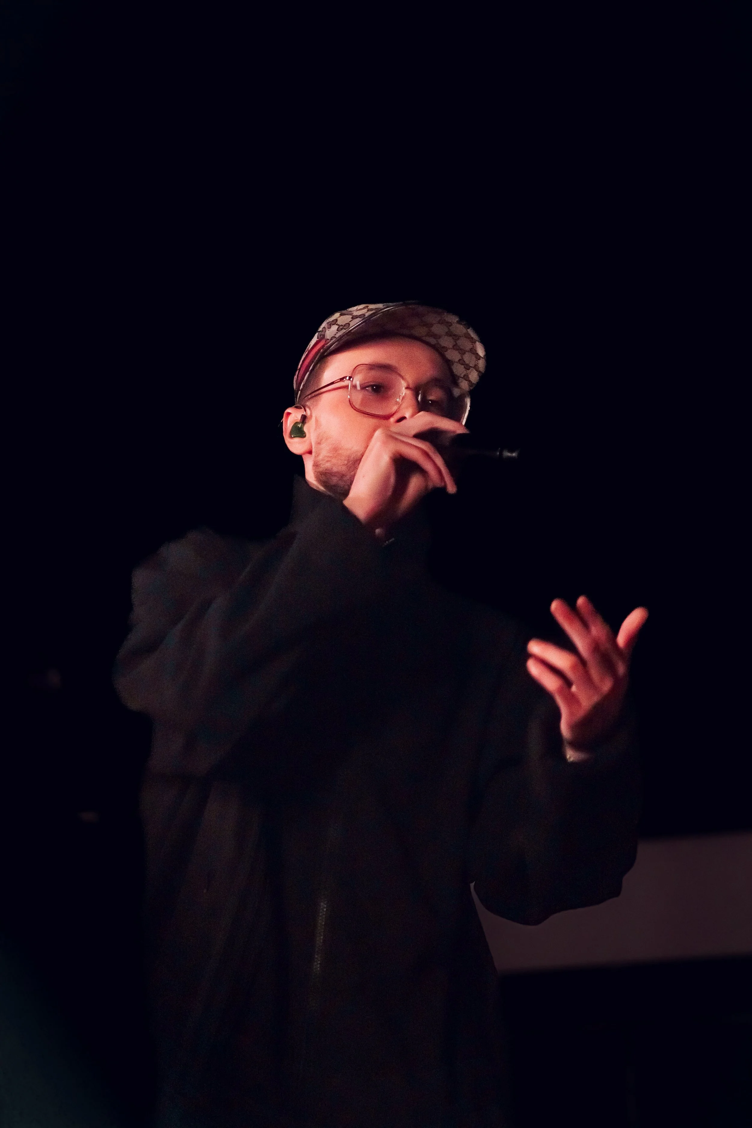 Jeune homme portant des lunettes, un chapeau à logo, chantant ou parlant en micro dans un environnement sombre.