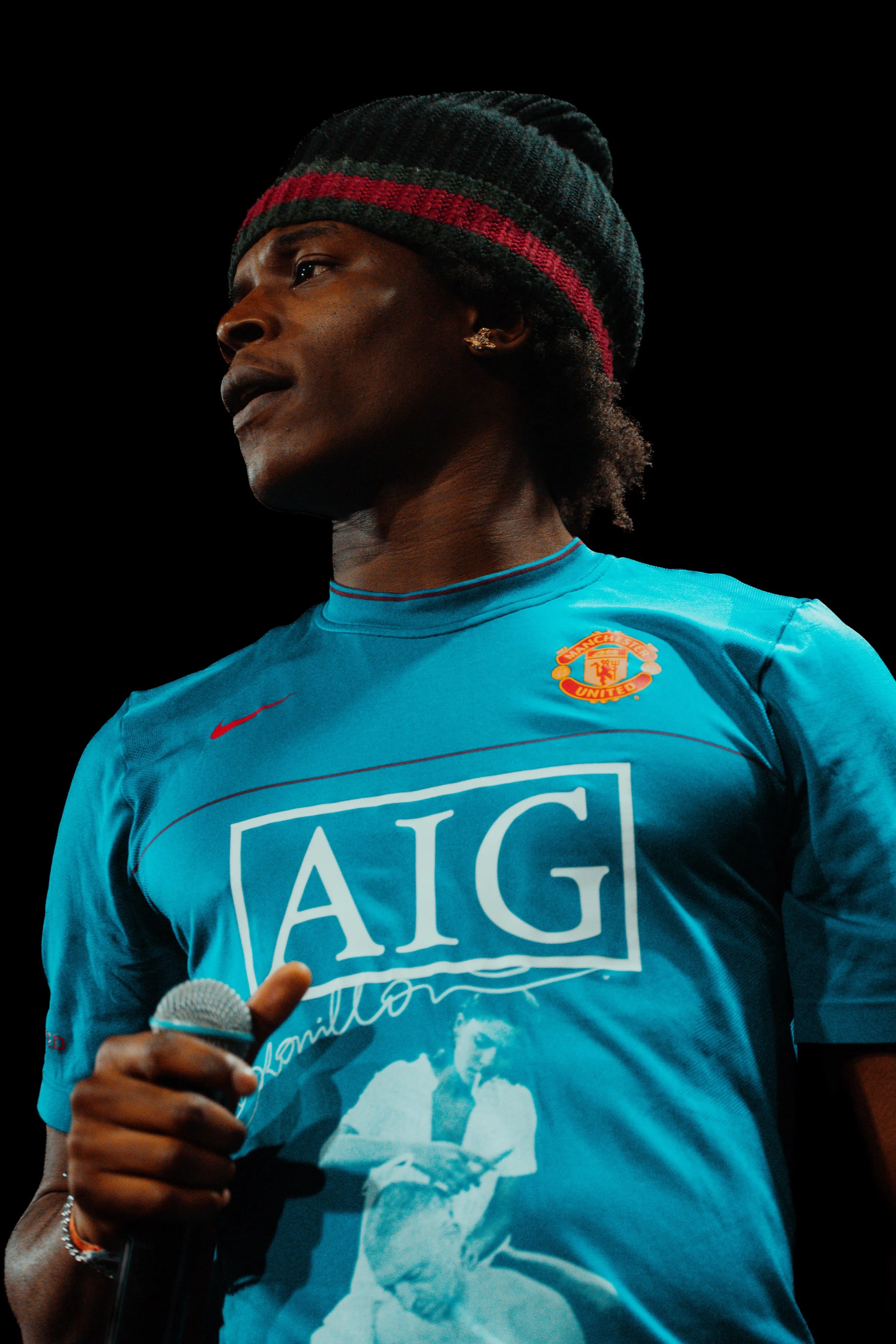 Un homme porte un maillot de football de Manchester United et un bonnet à rayures rouges et noires, tenant un microphone et regardant vers le haut.