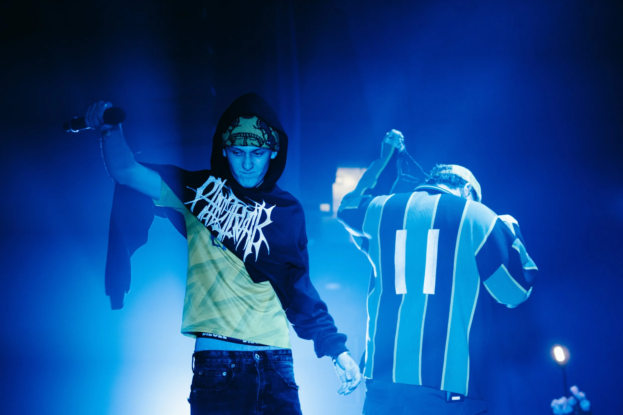 Deux jeunes hommes en hoodie sur scène, un avec un micro dans la main, éclairage bleu, ambiance de concert