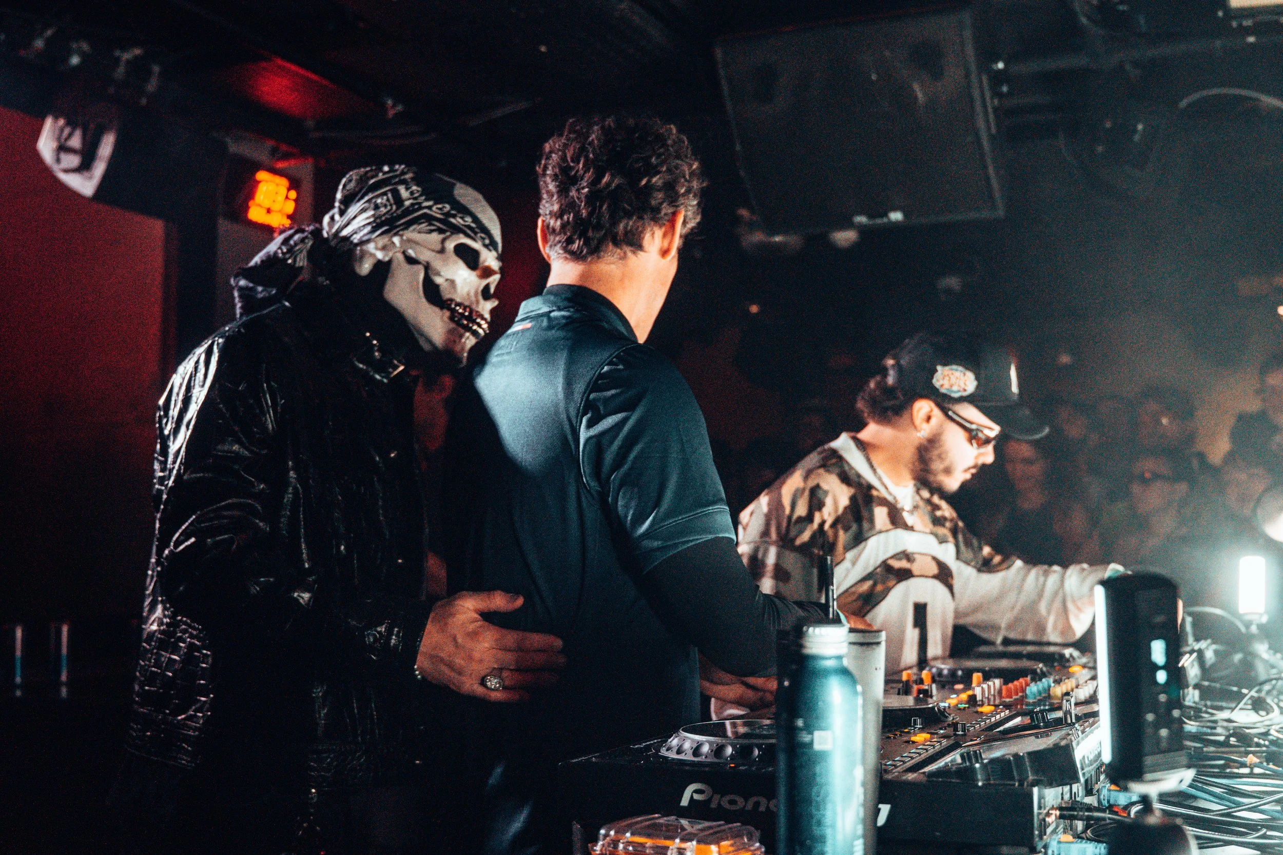Trois DJ sur scène, l'un porte un masque de squelette, un autre porte une casquette de baseball, la scène est éclairée par une lumière brillante.