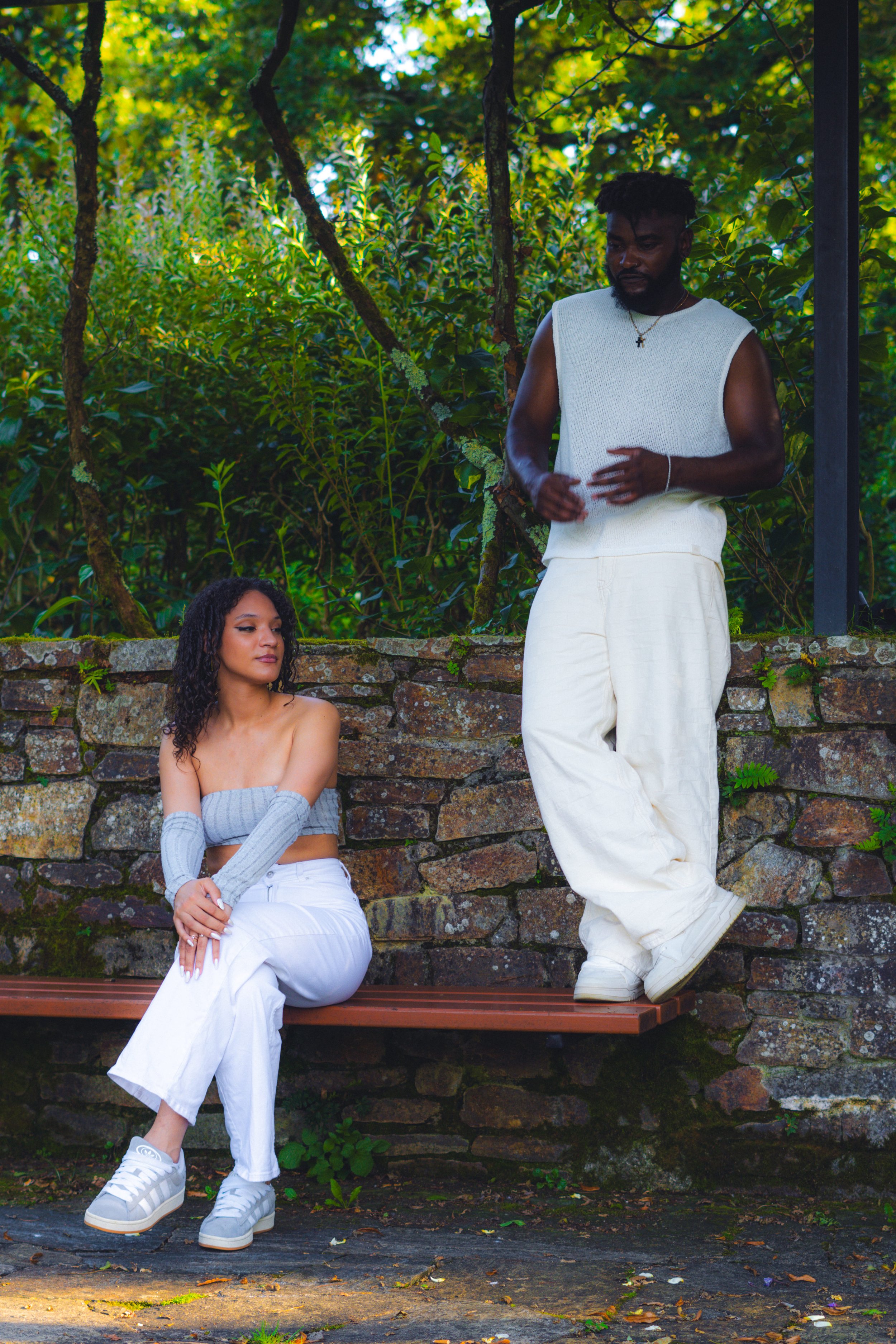 Une femme assise sur un banc en bois, portant un haut gris et un pantalon blanc, et un homme debout à côté, portant un haut sans manches blanc et un pantalon blanc, dans un environnement extérieur avec des arbres verts et une vieille muraille.