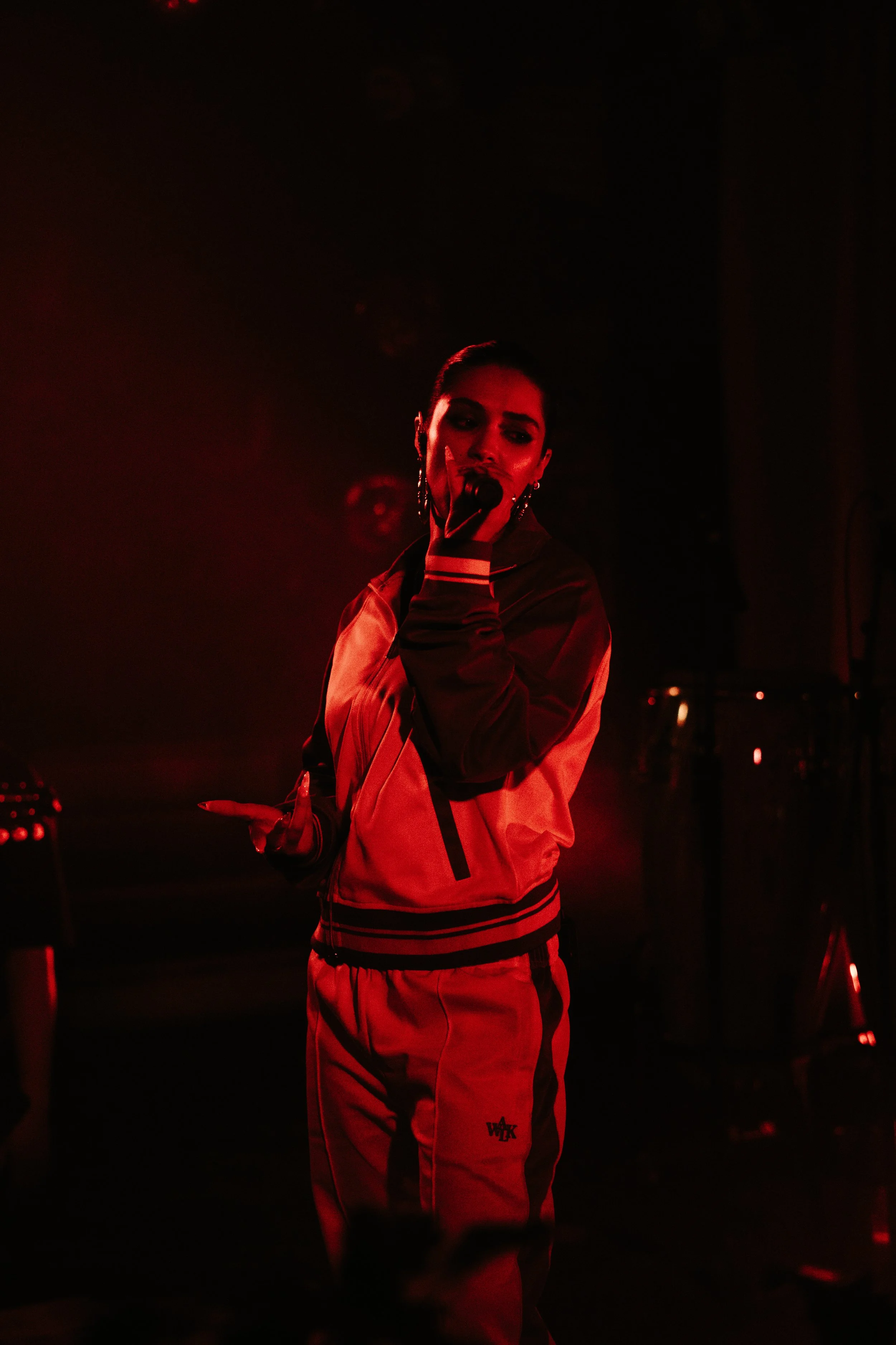 Une femme chanteuse sur scène, portant un survêtement rouge et noir, en train de chanter dans une lumière rougeâtre.