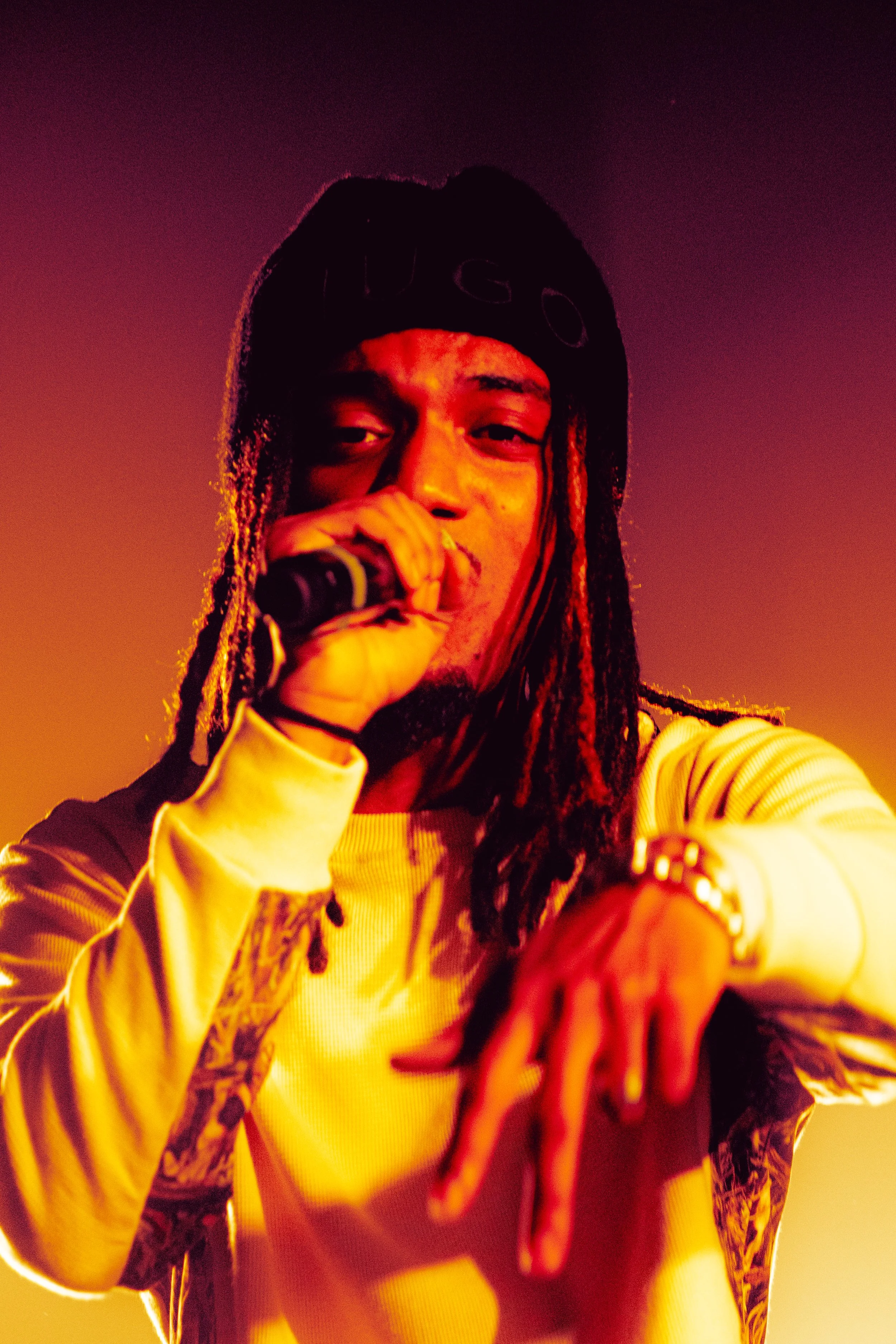 Un rappeur avec des dreadlocks, portant un bonnet noir avec le texte 'HUGO', chante en tenant un micro, dans un éclairage orange et jaune.