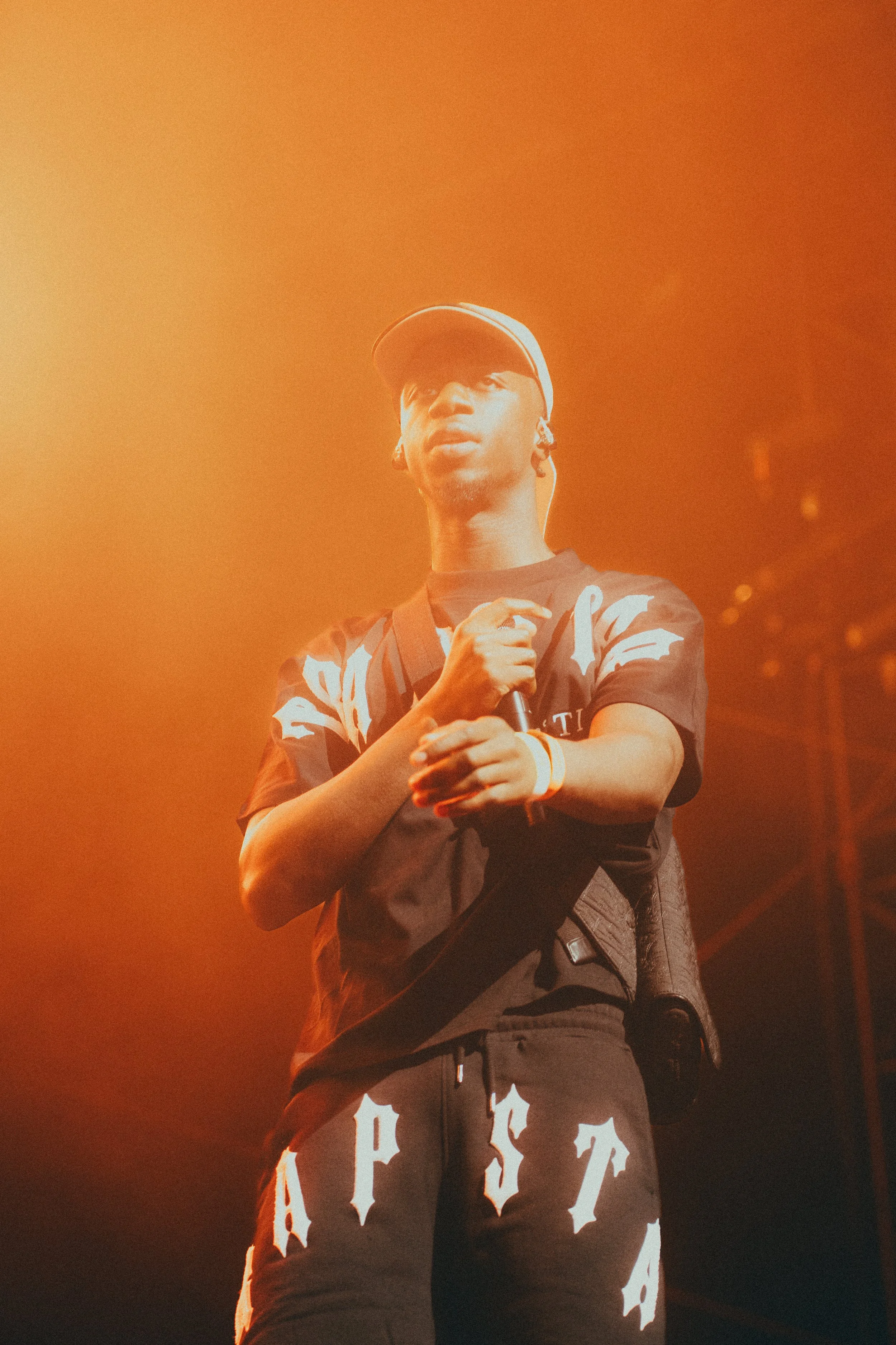Un jeune homme sur scène, portant une casquette, un t-shirt noir avec des motifs blancs, et un pantalon noir avec des lettres blanches, tenant un microphone, sous un éclairage orange intense.