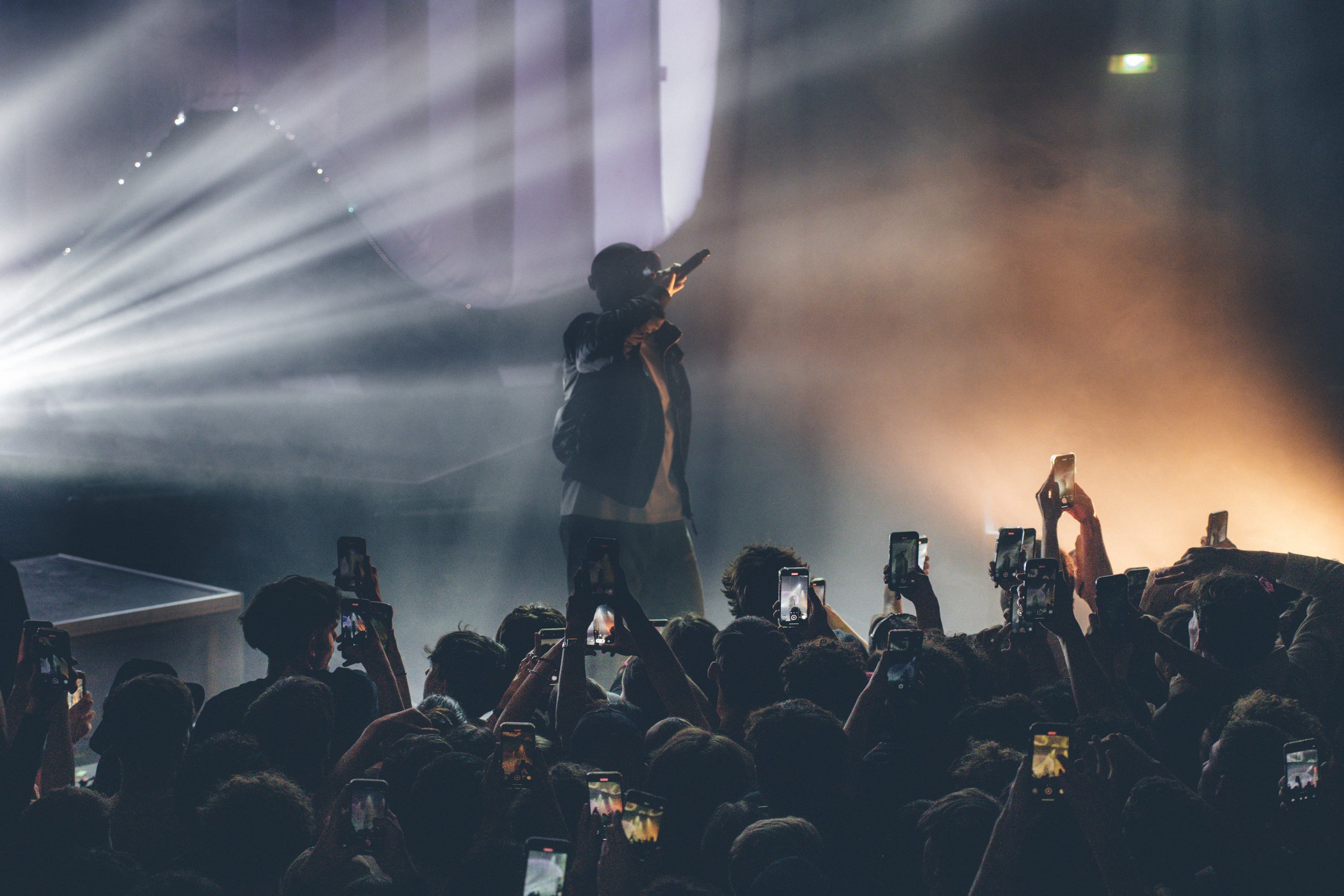Un artiste chante sur scène pendant un concert, entouré de nombreux spectateurs qui prennent des photos avec leurs téléphones.