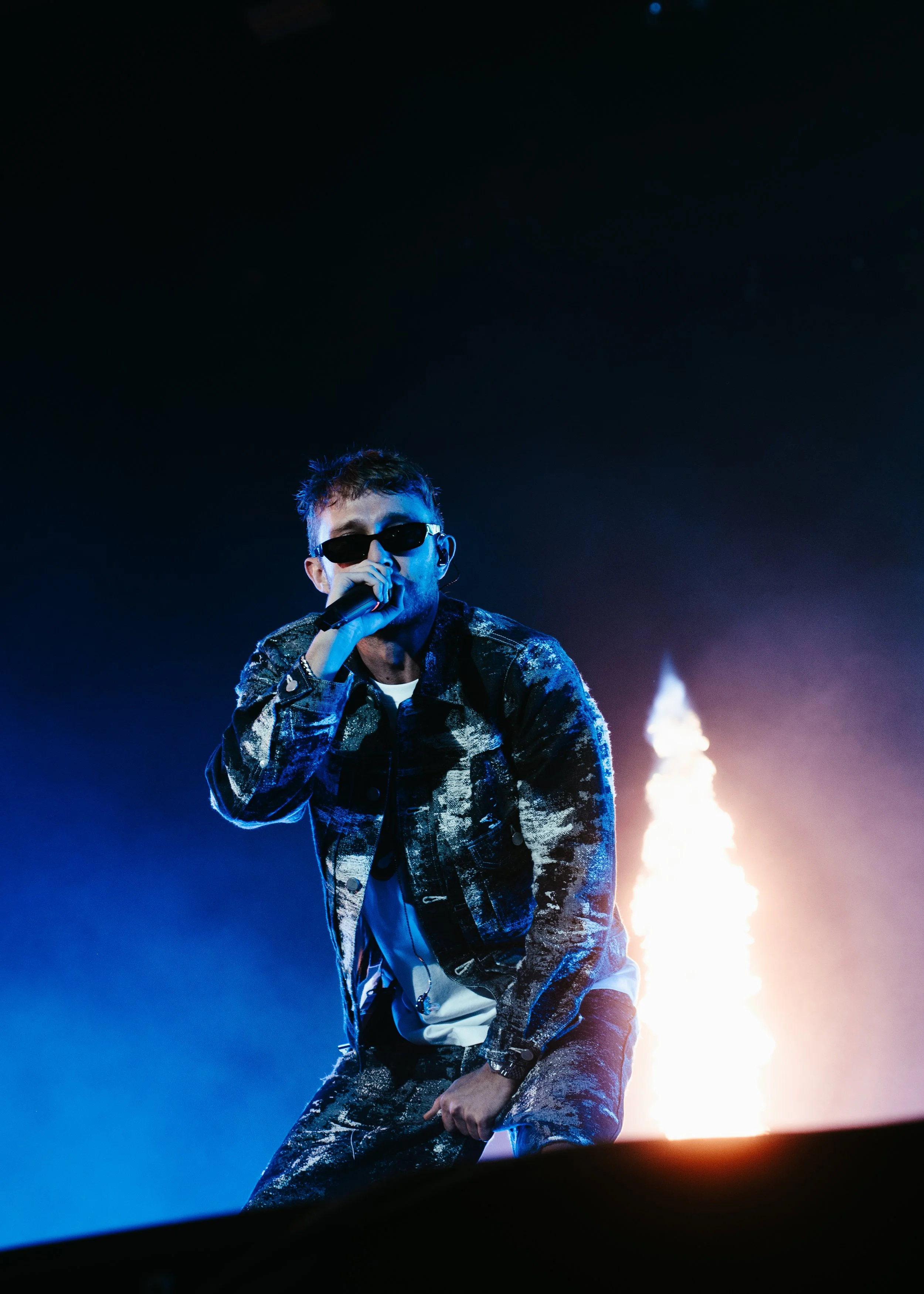 Un chanteur portant des lunettes noires et un vêtement à motifs, tenant un microphone sur scène, avec un feu d'artifice en arrière-plan.