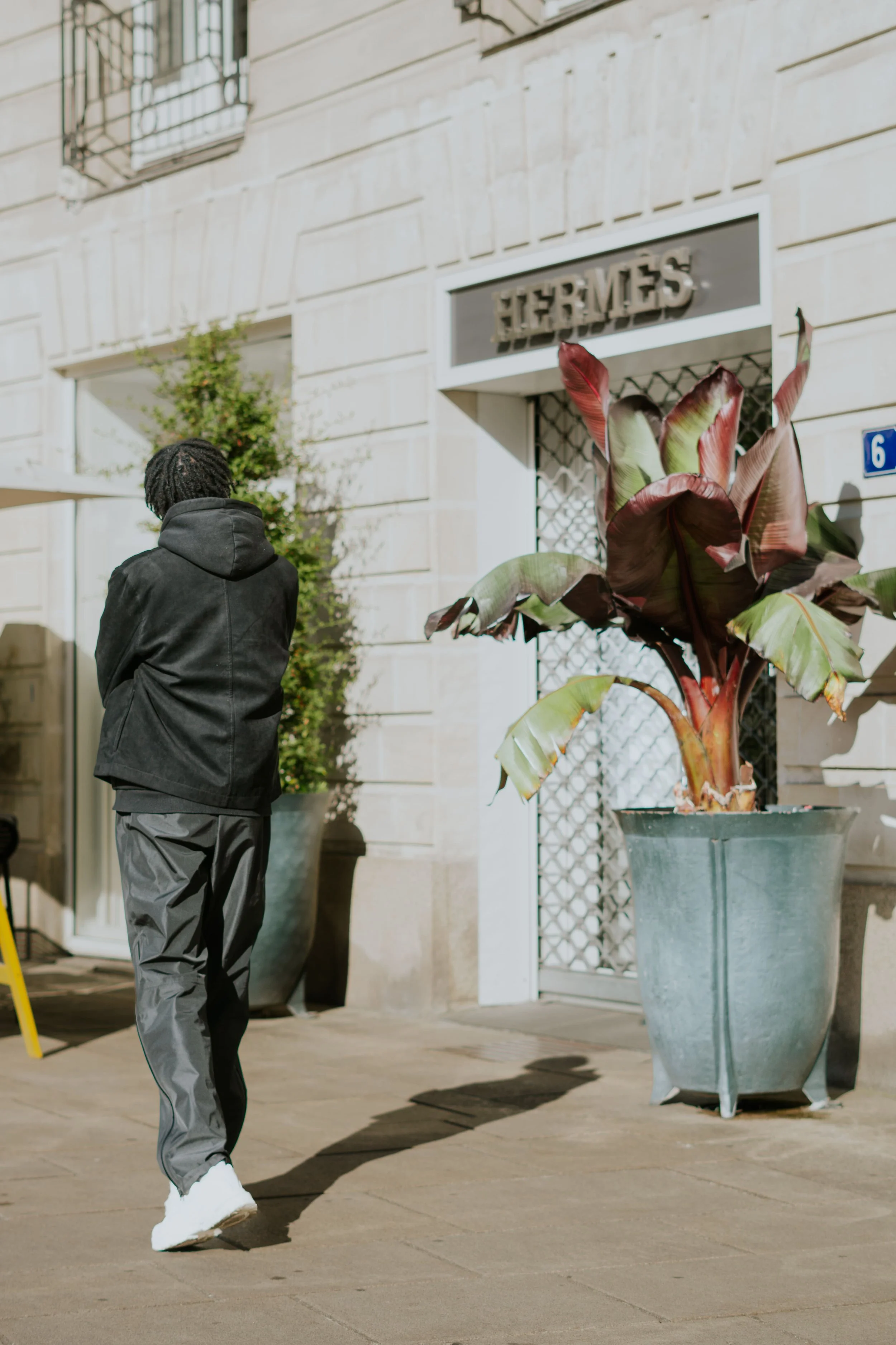 Un homme marche devant une boutique Hermès, avec deux grands pots de plantes devant la façade d'un bâtiment en pierre
