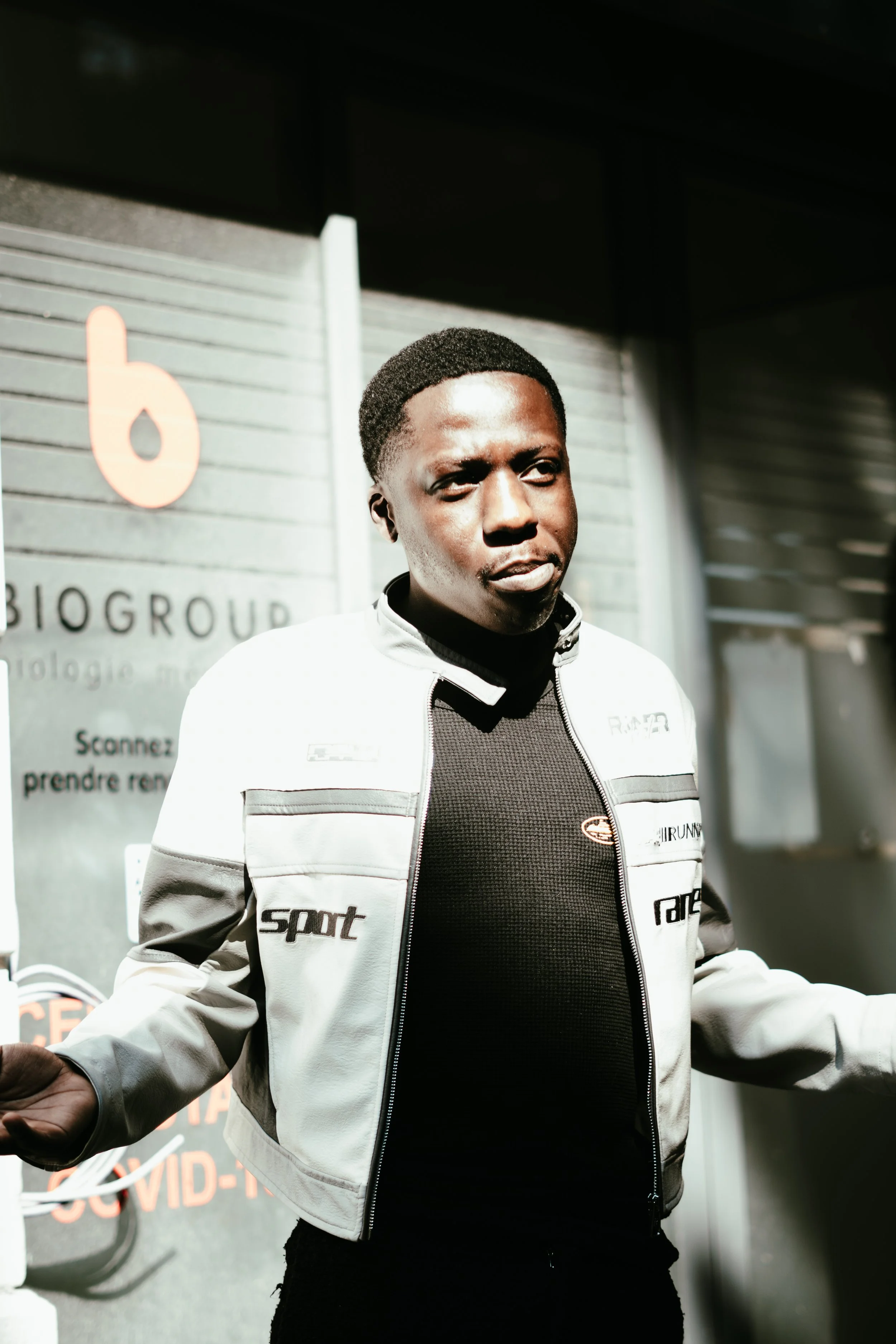 Un homme porte une veste de motard en cuir blanc avec des inscriptions noires sur fond sombre, debout à l'extérieur devant un panneau publicitaire.